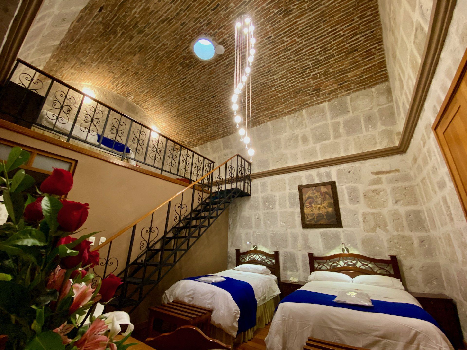 Dormitorio con paredes de piedra, dos camas, escalera a un loft y una lámpara colgante.