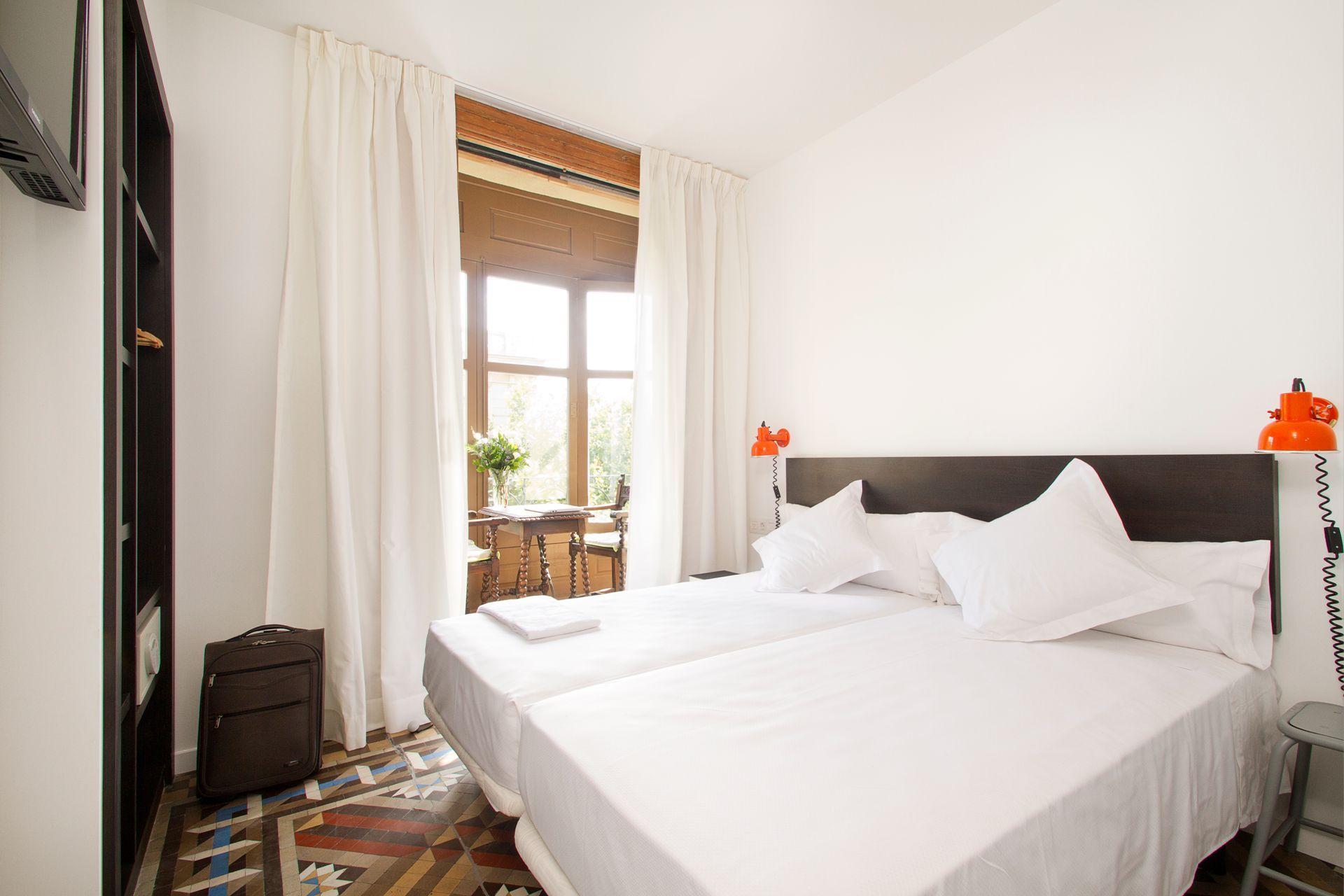 Habitación luminosa con ropa de cama blanca, cabecero oscuro y ventana con cortinas y vistas al balcón.