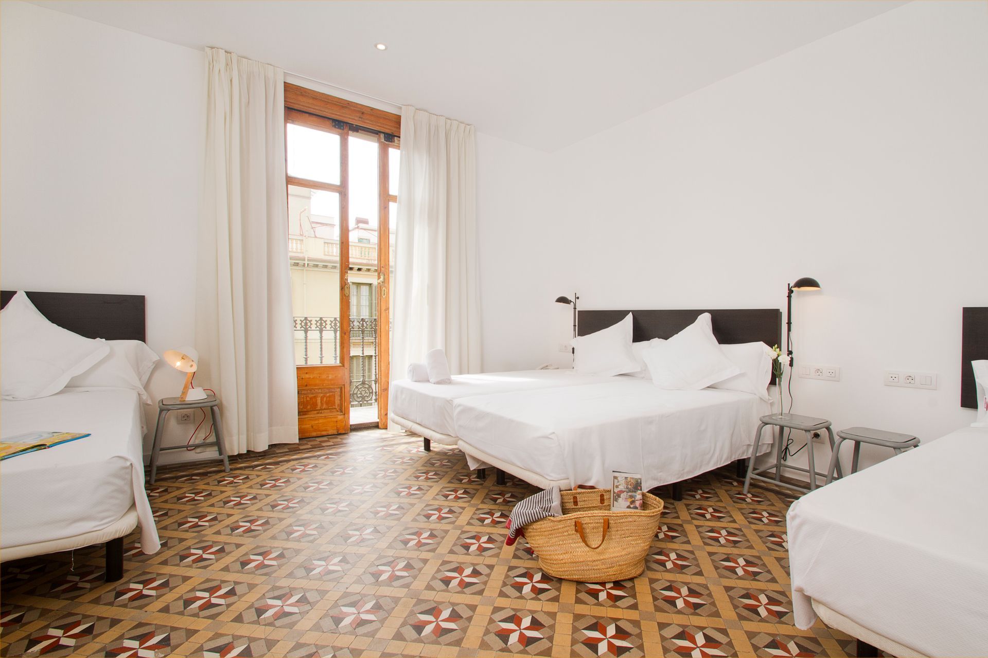 Habitación luminosa con tres camas blancas, suelo de baldosas estampadas y puertas de balcón con cortinas transparentes.