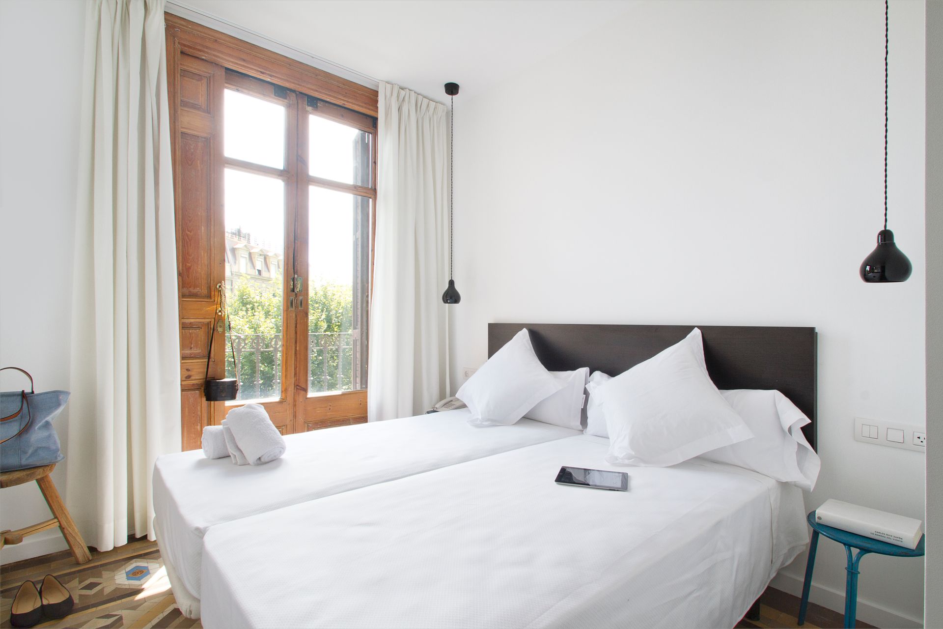 Habitación luminosa con sábanas blancas, ventana con marco de madera y lámparas colgantes negras.