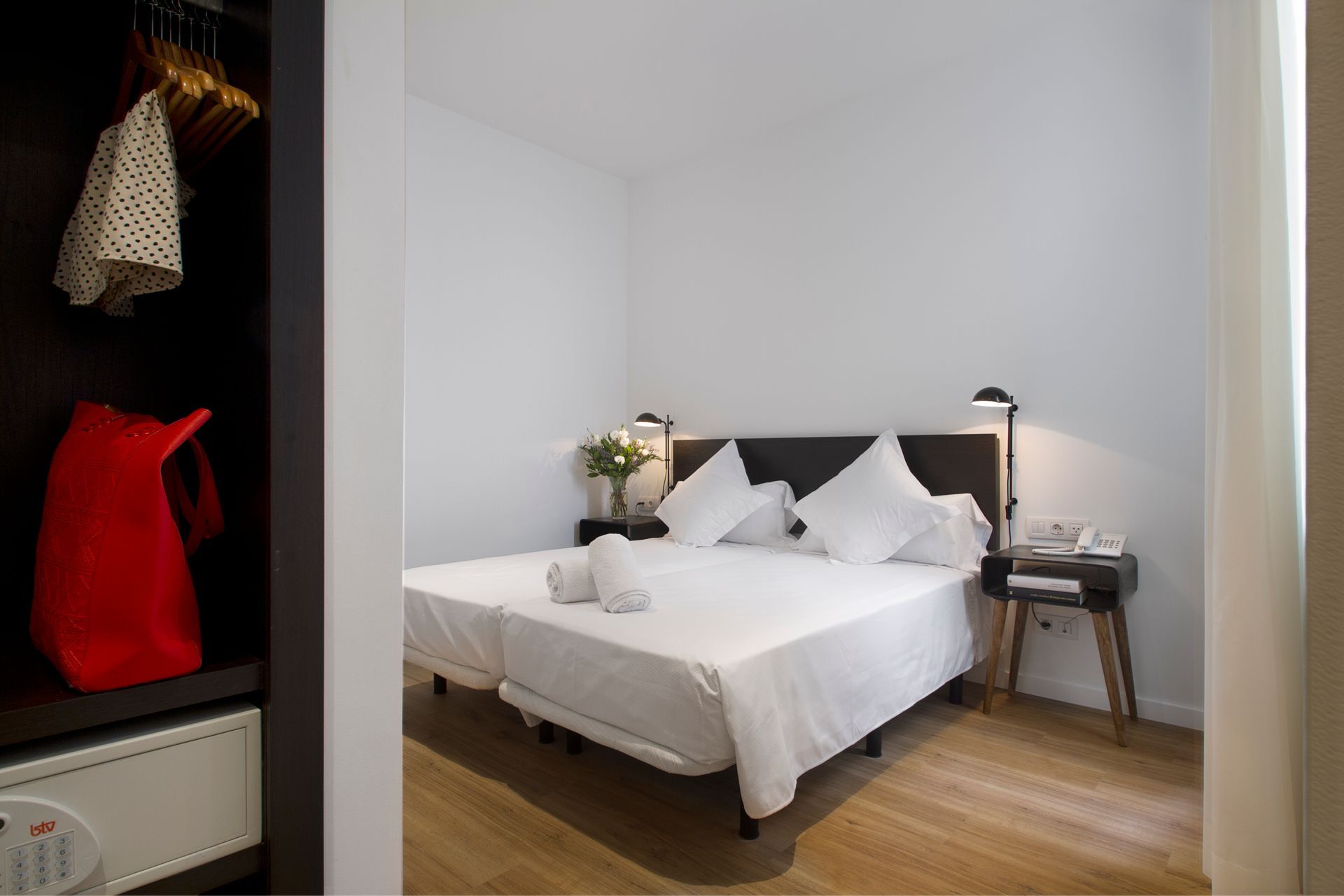 Habitación minimalista estilo hotel con cama blanca, cabecero negro, suelo de madera y lámpara de noche.