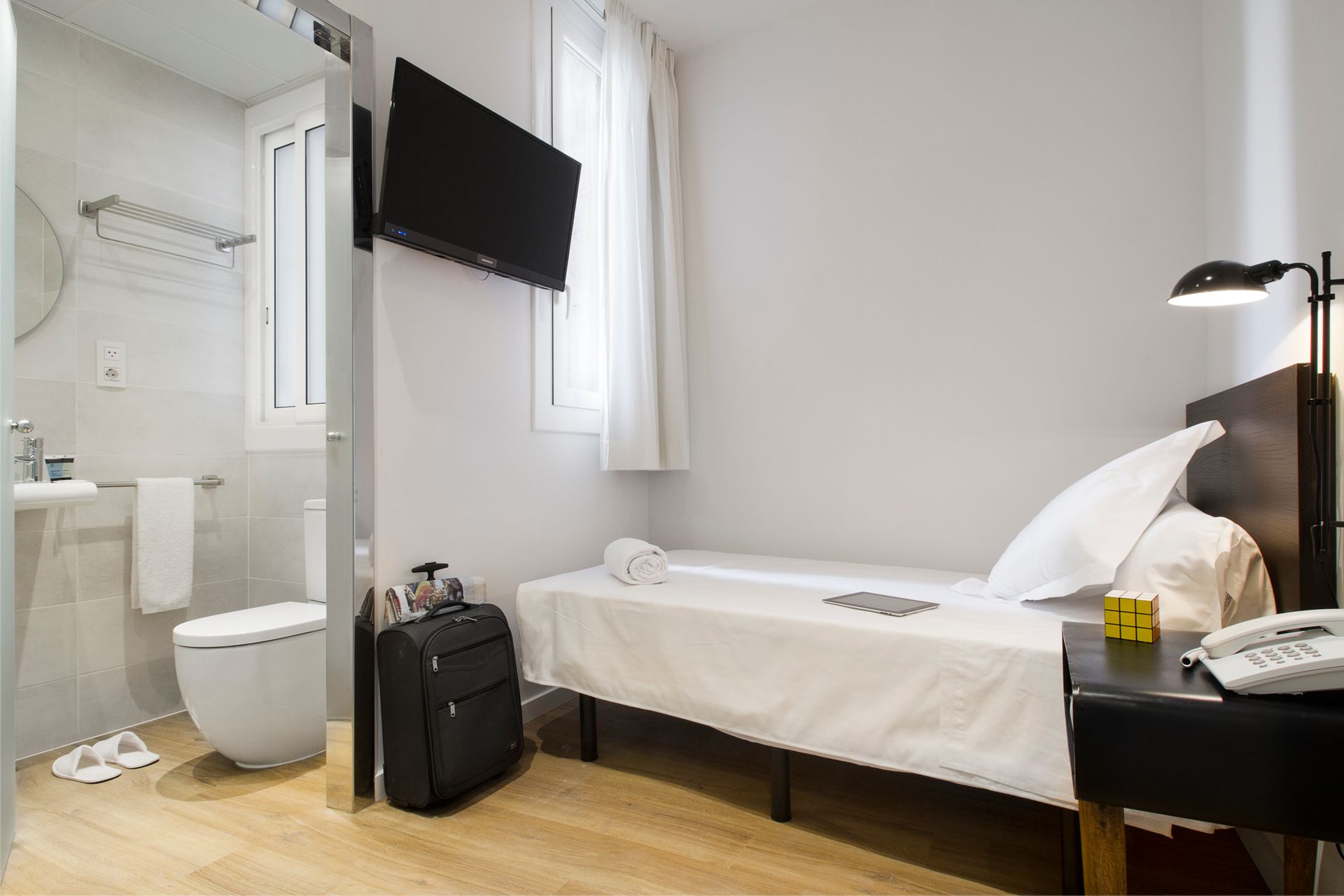 Habitación de hotel luminosa con cama individual, televisor de pared, escritorio y baño abierto.