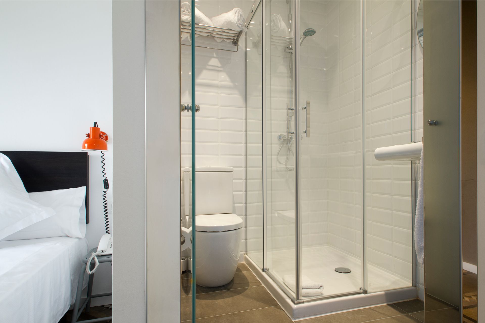 Baño moderno con ducha de cristal, inodoro y cama visibles al lado.