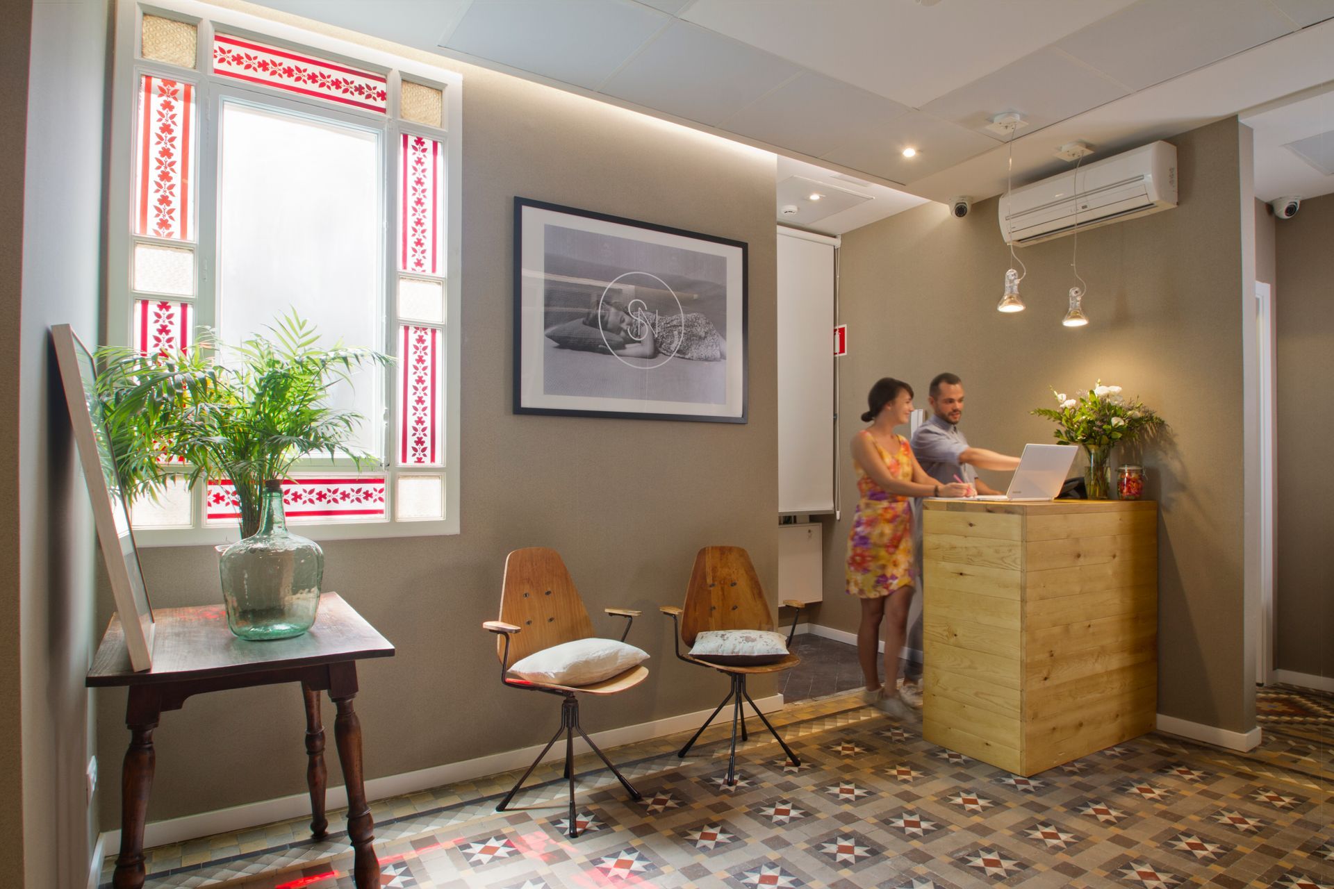 Área de recepción luminosa con azulejos estampados, plantas, cuadros y dos personas en un mostrador de madera.