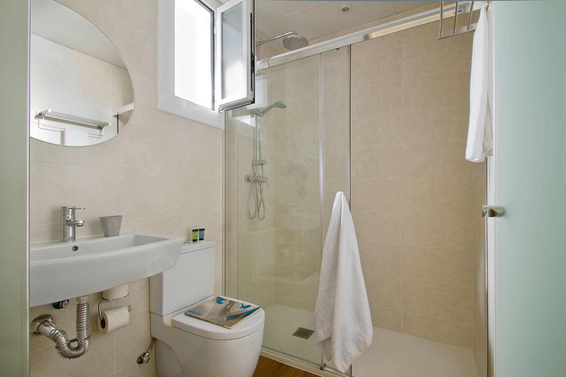 Baño pequeño con lavabo, inodoro, ducha, espejo y toallas blancas en un interior de color beige.