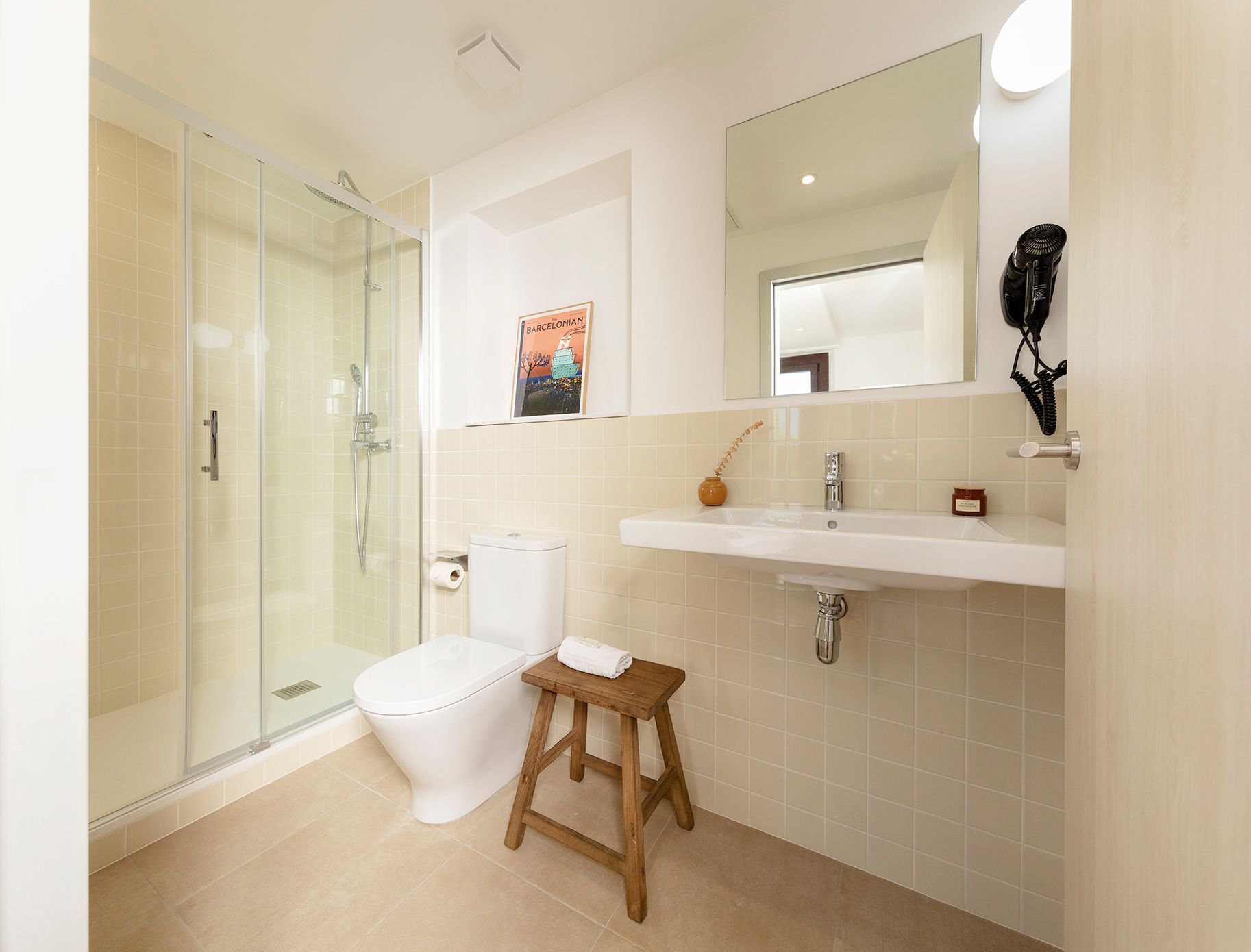 Baño moderno y luminoso con ducha de cristal, inodoro, lavabo con mueble, espejo y taburete de madera.