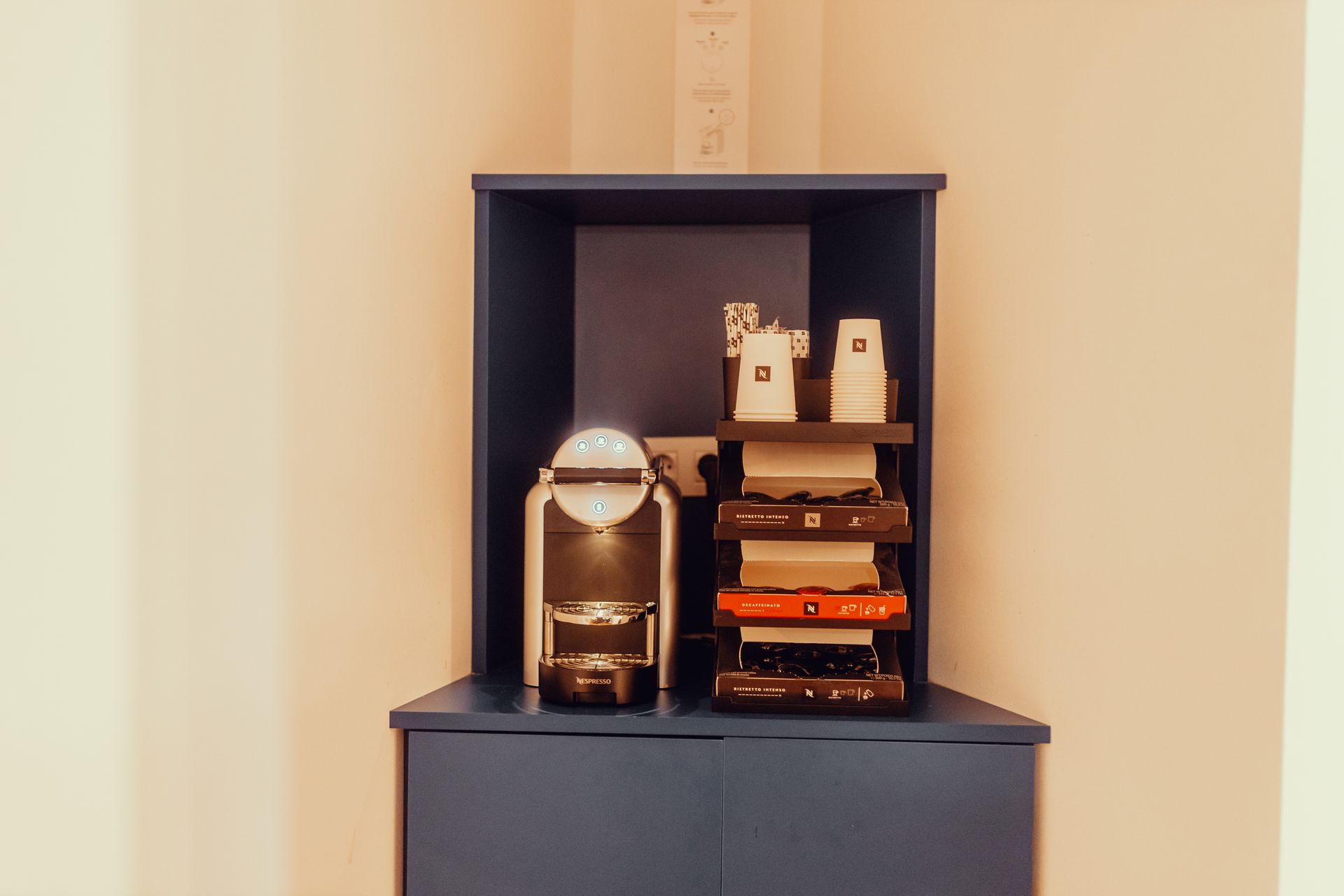 Pequeña zona de café con máquina, tazas y vasos apilados sobre un mueble oscuro contra una pared beige.