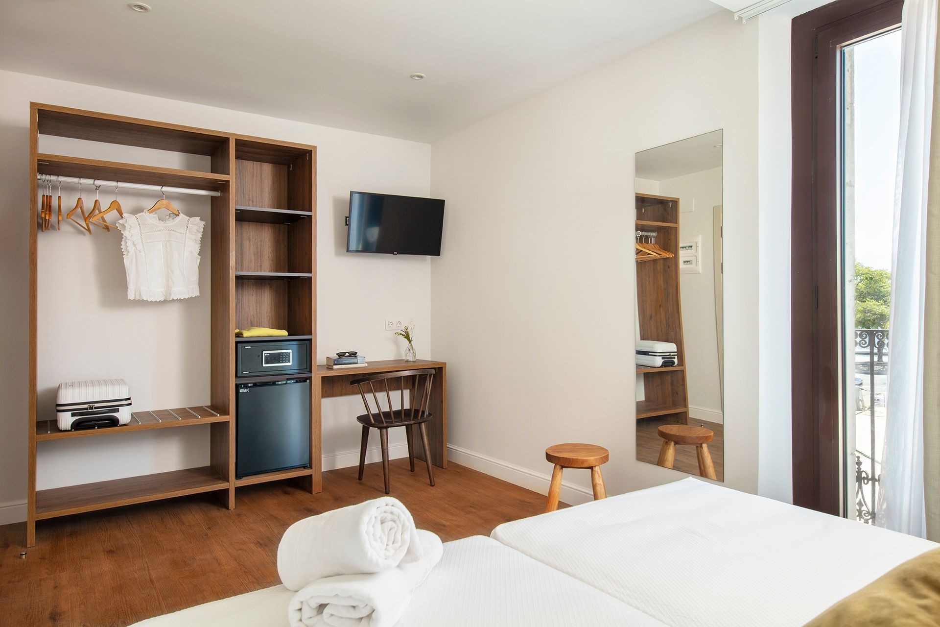 Habitación de hotel luminosa con armario abierto, escritorio, televisor de pared, cama y puerta que da al balcón.