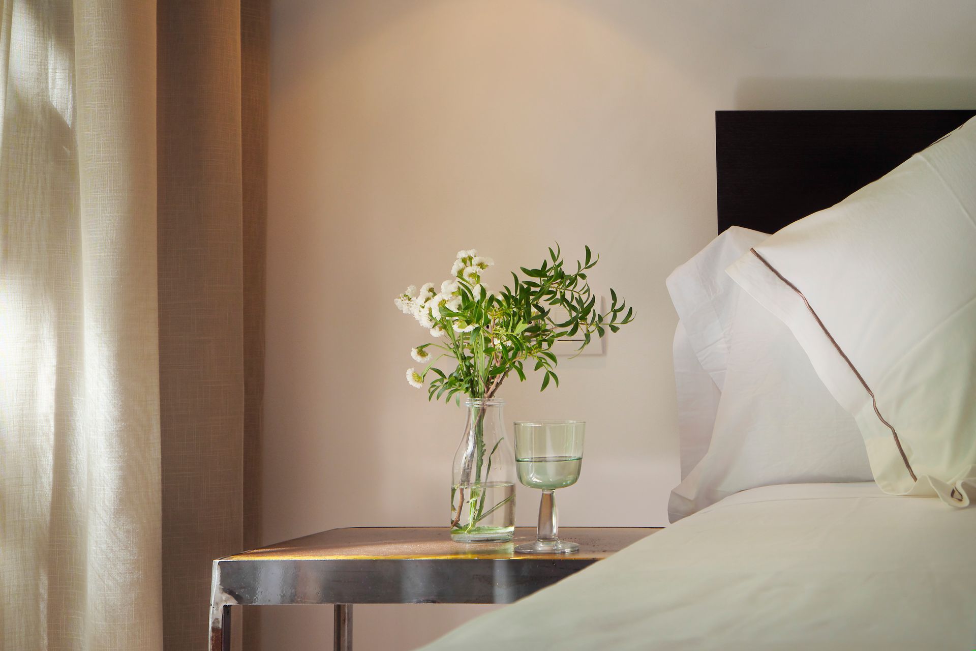 Una cama bien hecha junto a una mesita de noche con un jarrón de flores blancas junto a una ventana tenuemente iluminada.