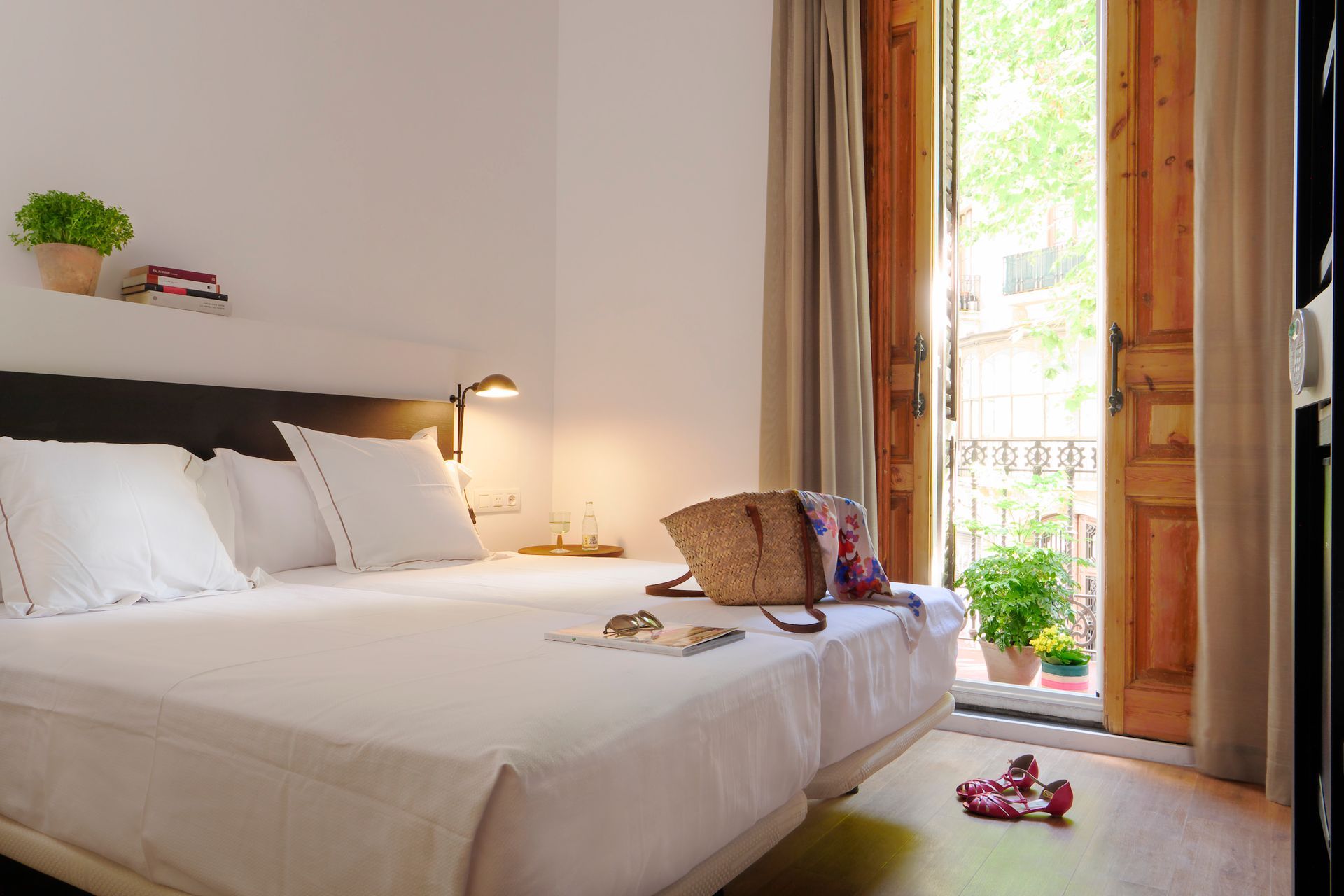 Habitación luminosa con cama, puerta de balcón abierta y luz natural, con un bolso y flores cerca de la ventana.