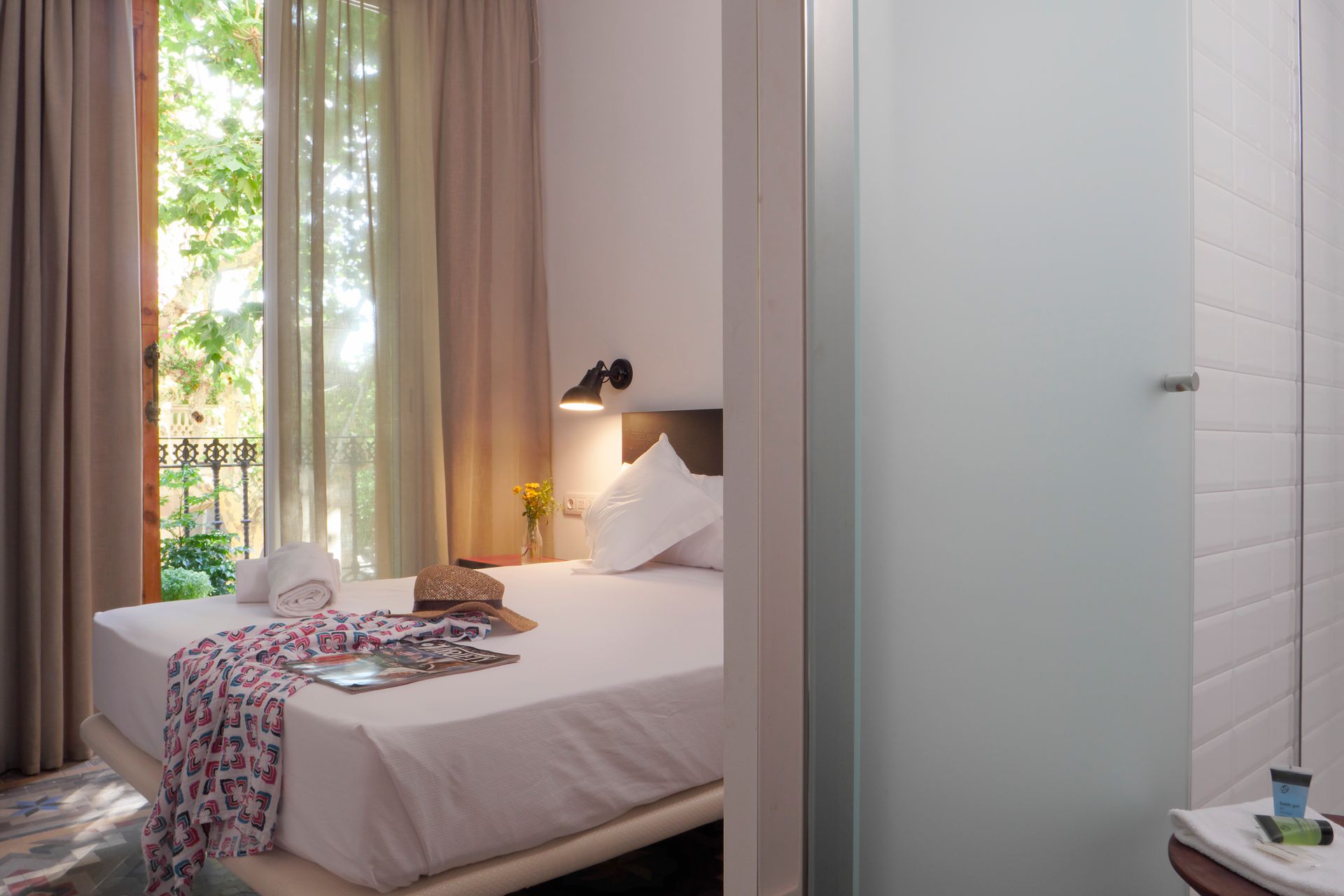 Habitación luminosa con una cama blanca sin hacer, lámpara de pared y puertas abiertas.