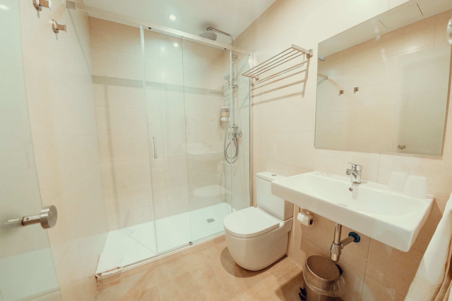 Baño luminoso con ducha de cristal, espejo de pared, lavabo blanco e inodoro sobre suelo de baldosas beige.