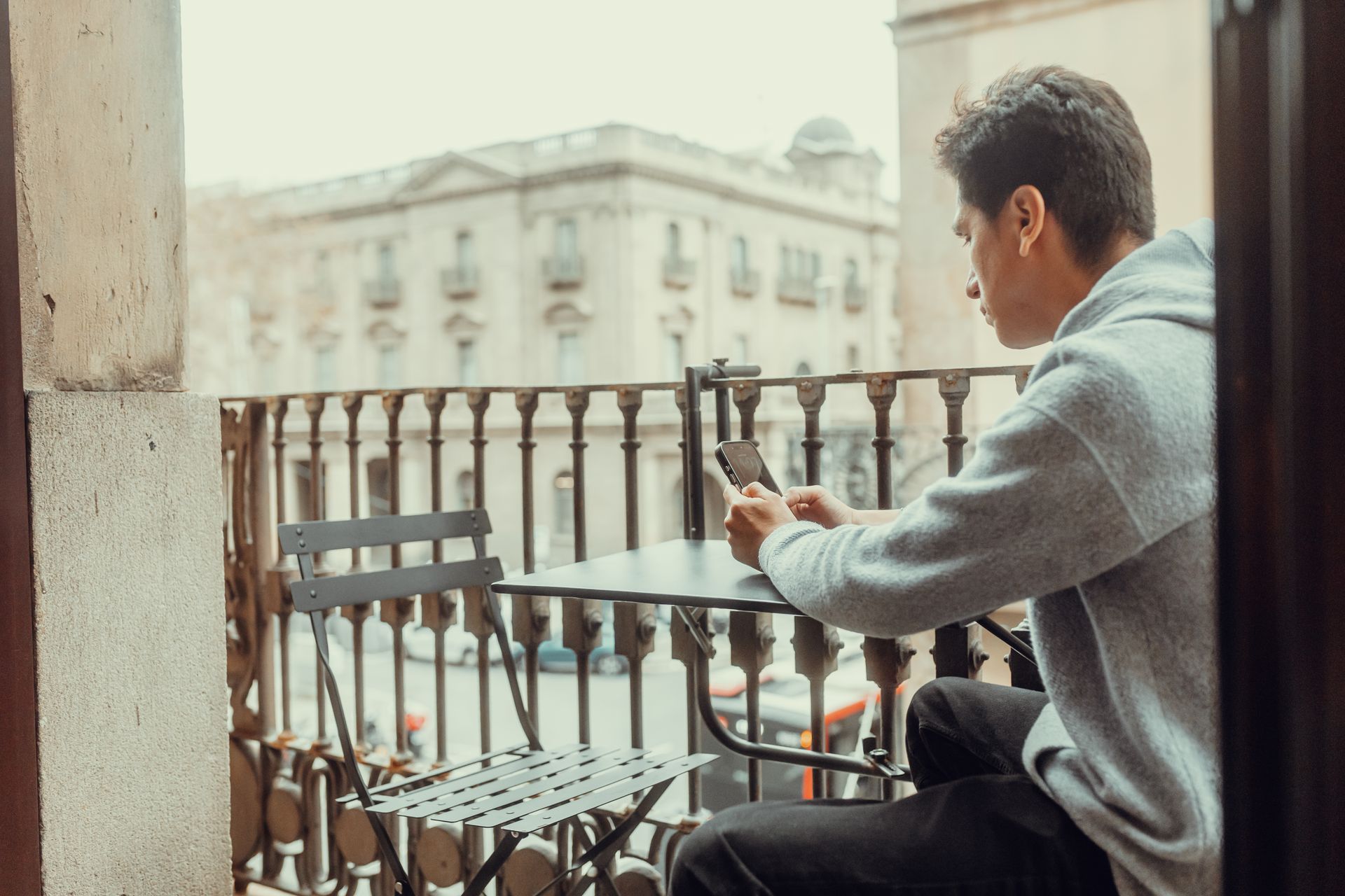 Persona sentada en un balcón usando un teléfono inteligente, con vistas a una calle de la ciudad y un edificio al fondo.