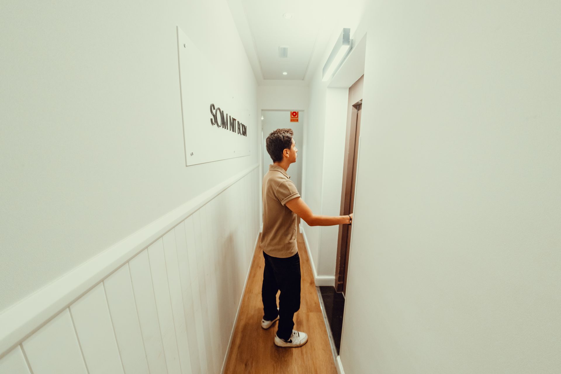 Una persona abre una puerta en un estrecho pasillo blanco con suelo de madera.