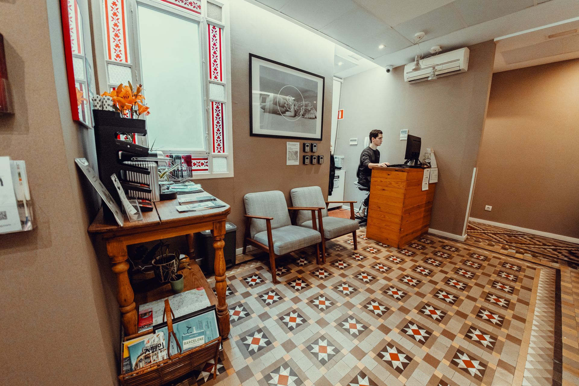 Pequeña área de recepción de oficina con piso de baldosas estampadas, sillas, cuadros enmarcados y un mostrador de madera.