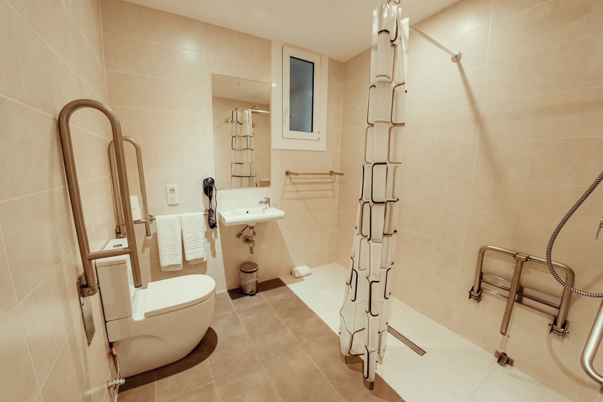 Baño accesible con inodoro, lavabo, ducha independiente y barras de apoyo; interior con azulejos de color beige.