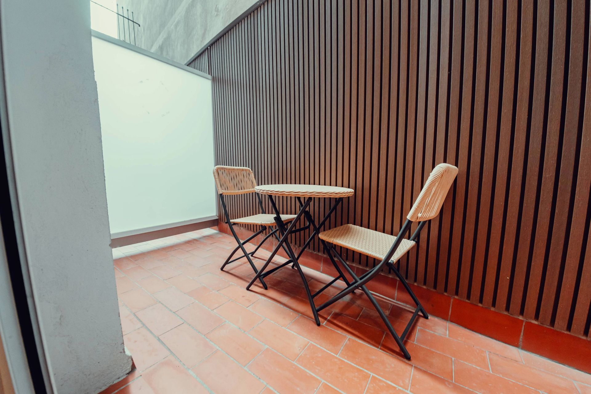 Pequeño patio con una mesa redonda y dos sillas plegables junto a una pared de listones de madera y una puerta de cristal.