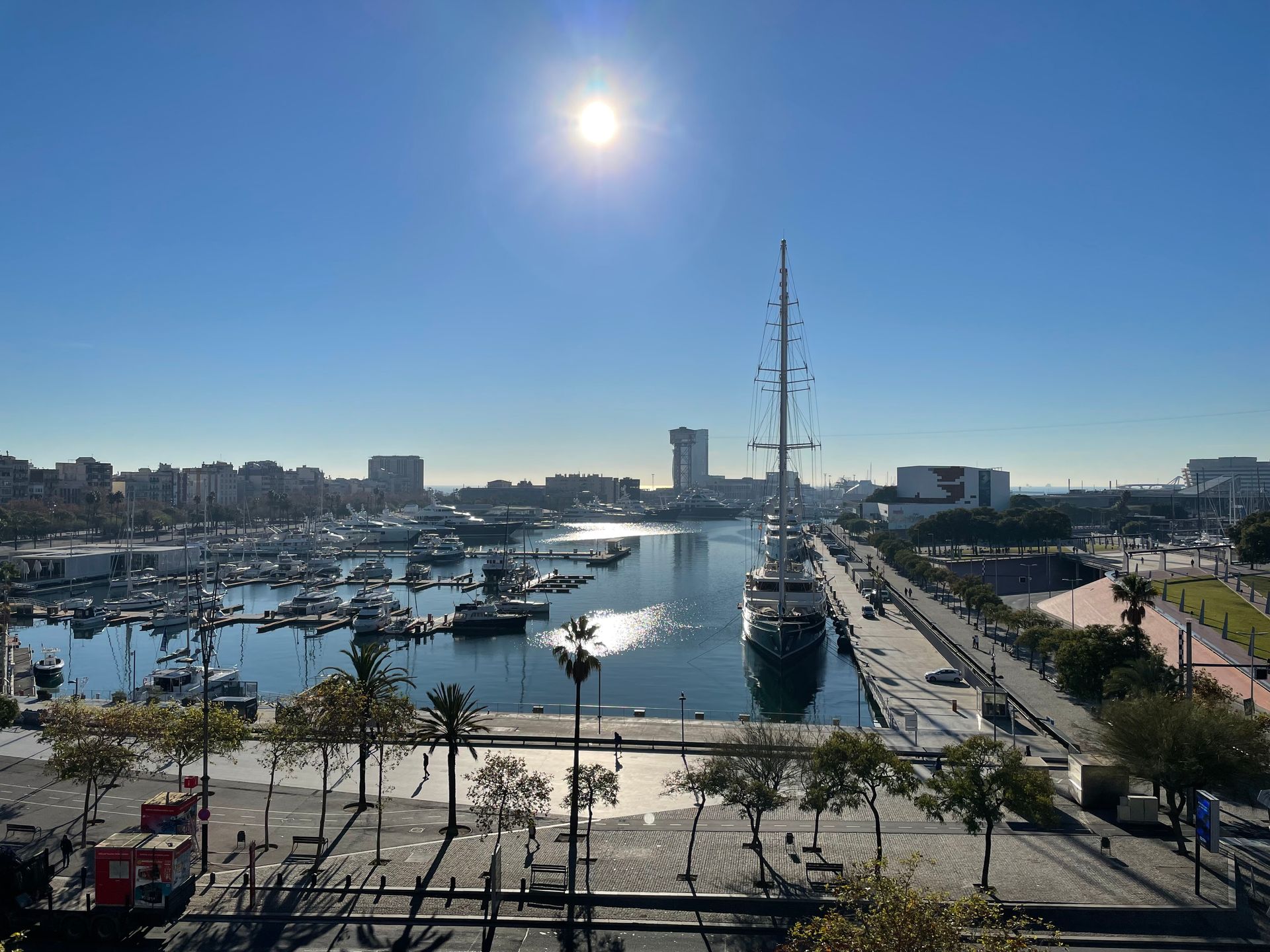 Un puerto deportivo soleado con barcos, un buque atracado y un mástil alto junto a un paseo marítimo.