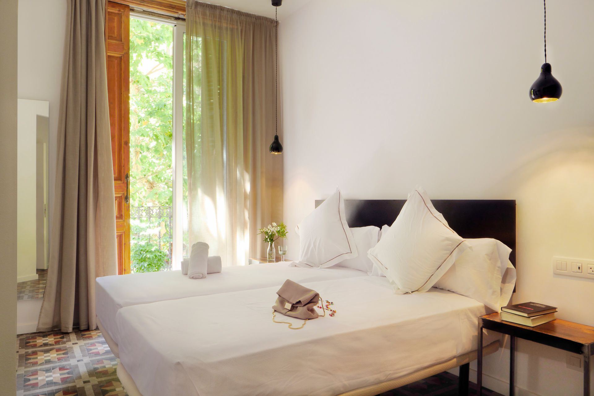 Habitación blanca luminosa y bien decorada, con una gran ventana, cortinas, mesita de noche y lámparas colgantes.