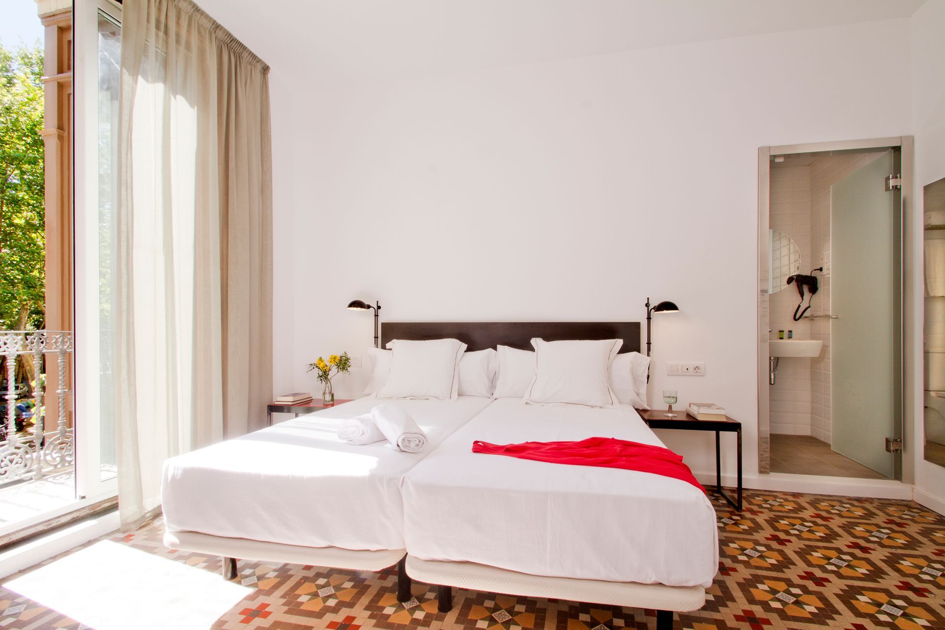 Habitación luminosa con dos camas blancas, manta roja, puertas que dan al balcón y suelo de baldosas estampadas.