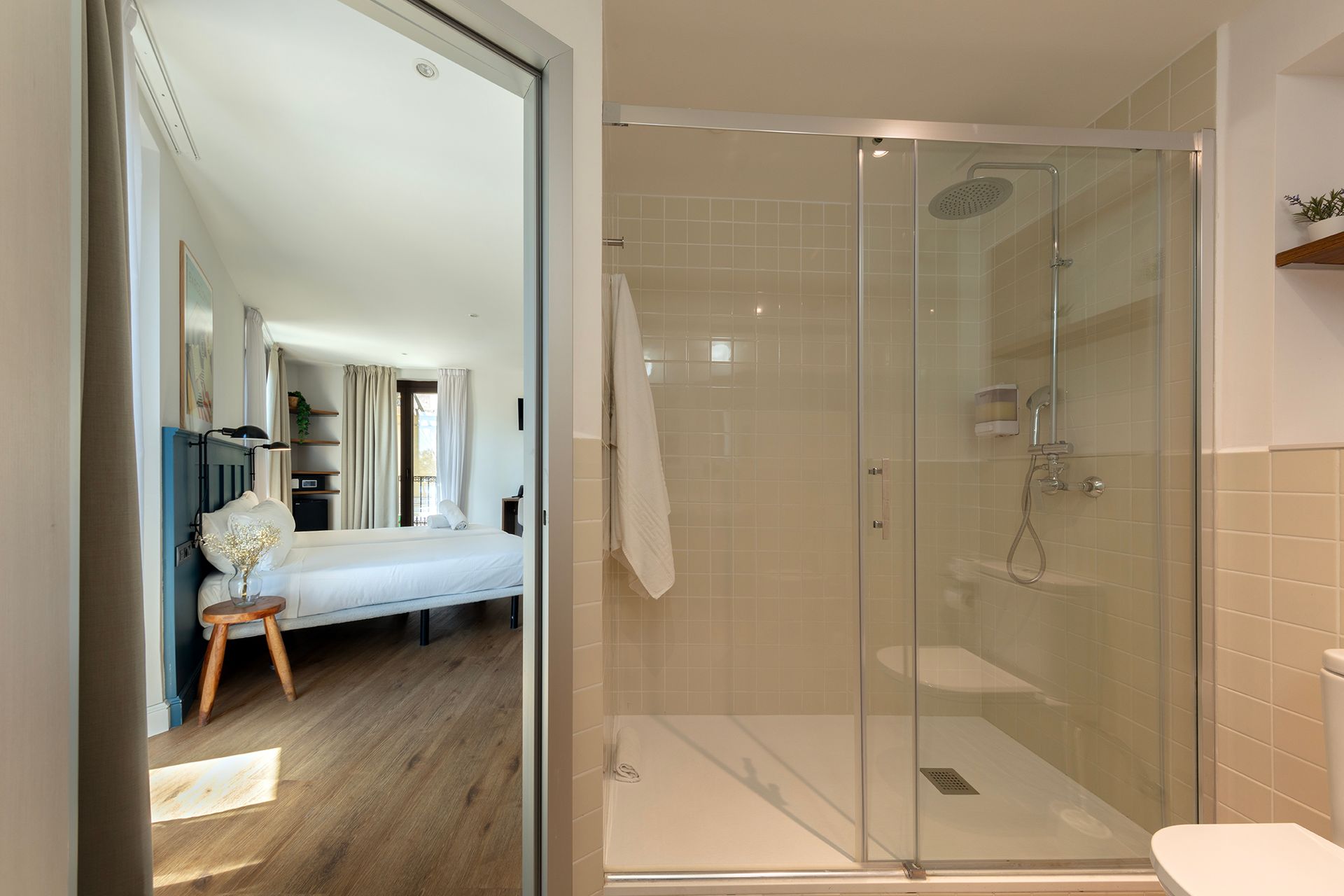 Baño moderno con ducha acristalada junto a un dormitorio visible a través de una puerta abierta.