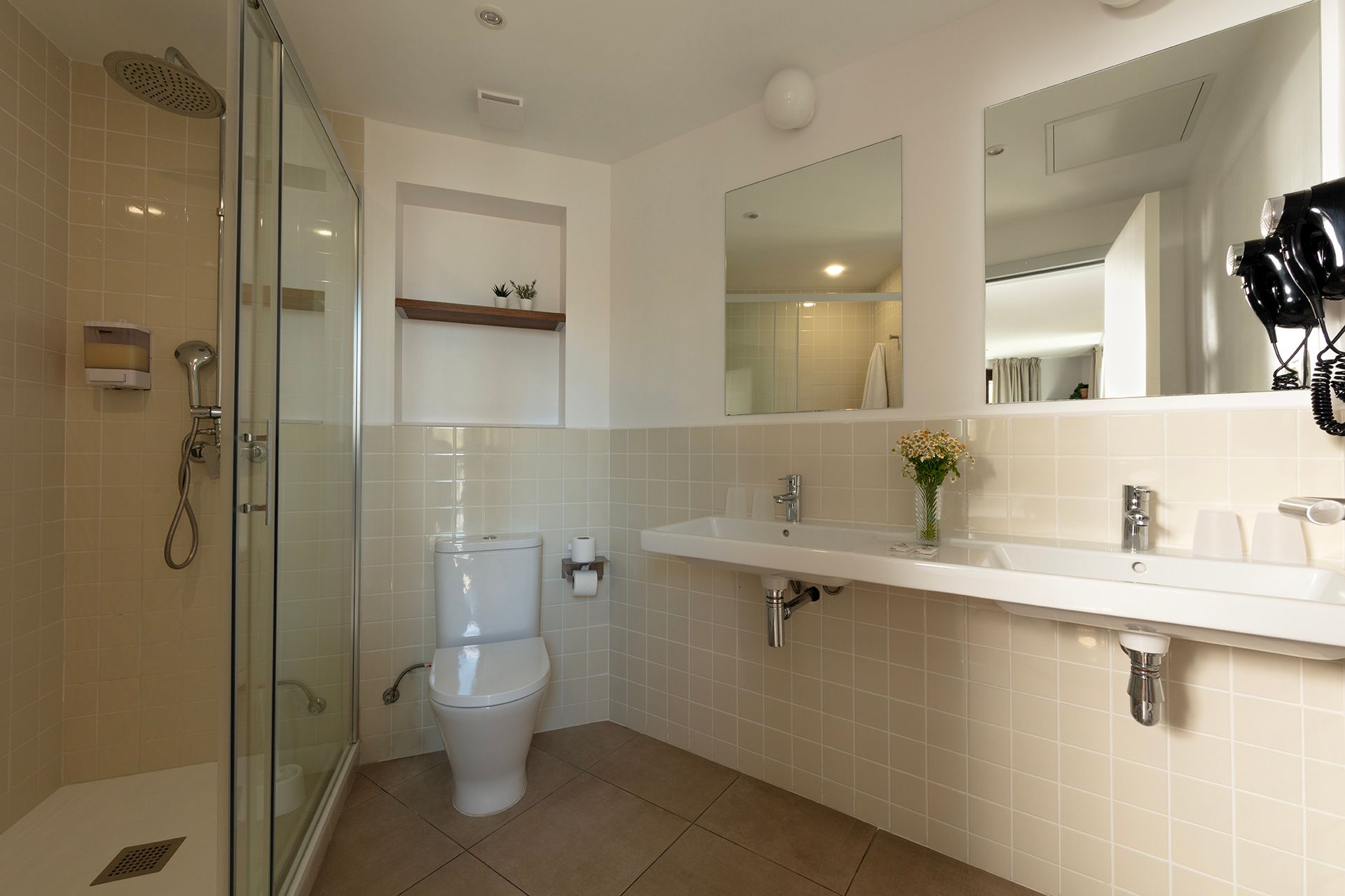 Baño moderno con inodoro, ducha, lavabo largo y espejo grande; azulejos beige y paredes blancas.
