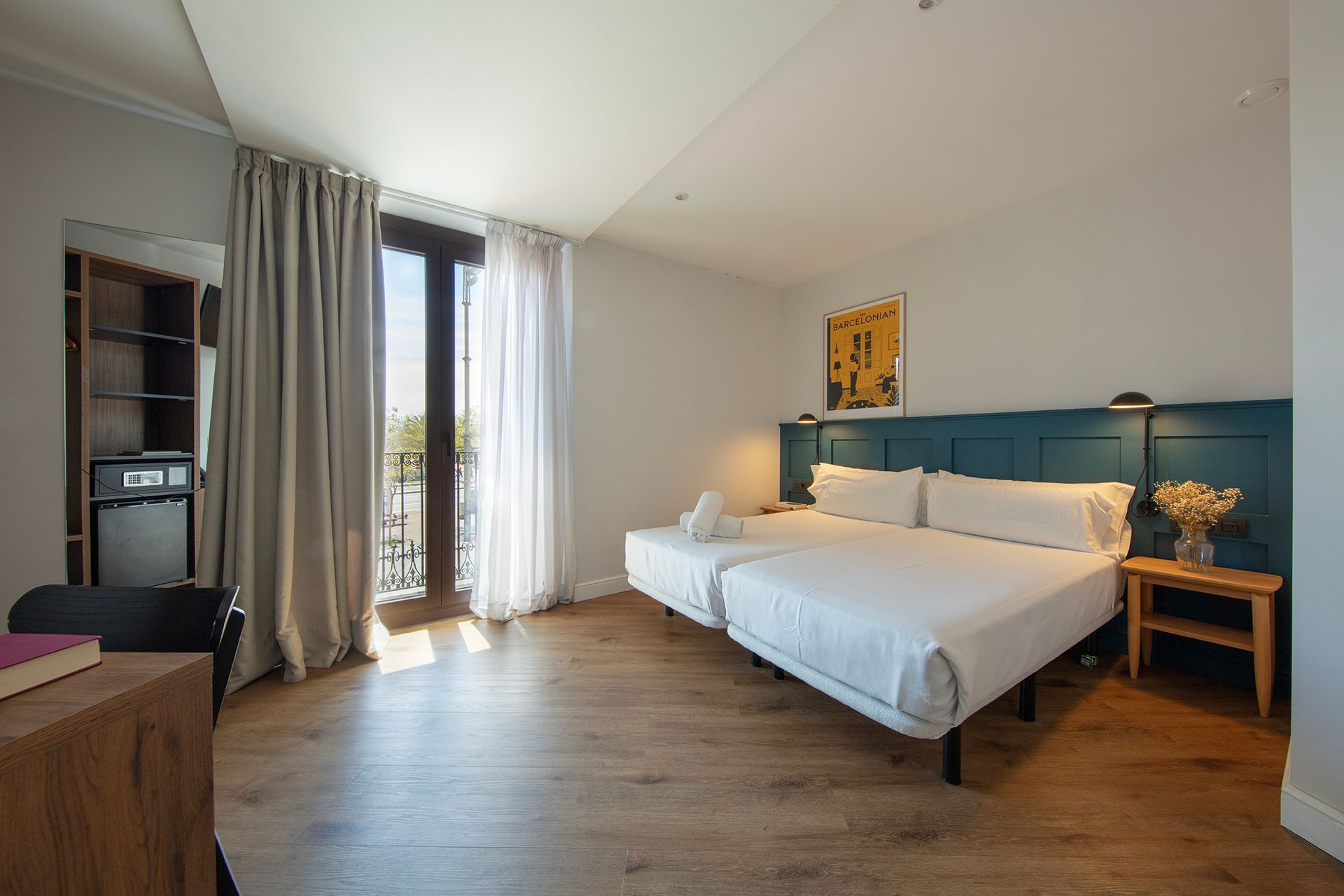 Dormitorio moderno con cabecero azul, ropa de cama blanca, suelo de madera y puertas de balcón con cortinas.