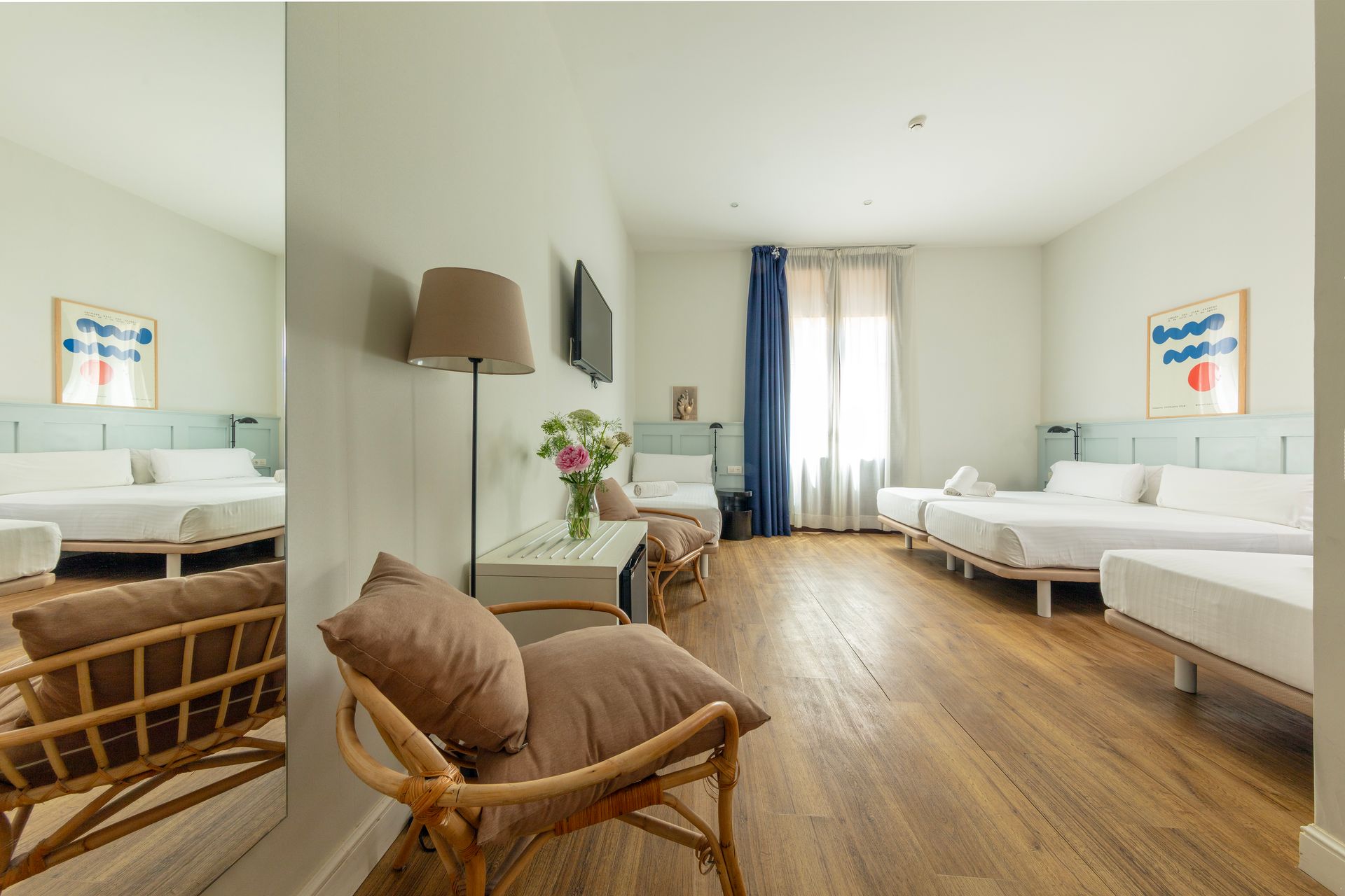 Habitación doble luminosa con silla de mimbre, camas blancas, suelos de madera y cortinas en las ventanas.