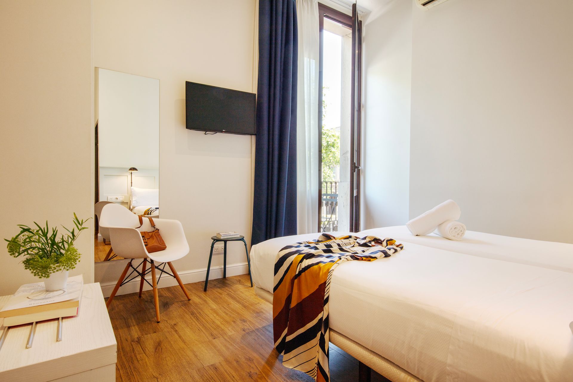 Dormitorio minimalista con cama blanca, suelo de madera, televisor montado en la pared, silla y puerta de balcón abierta.