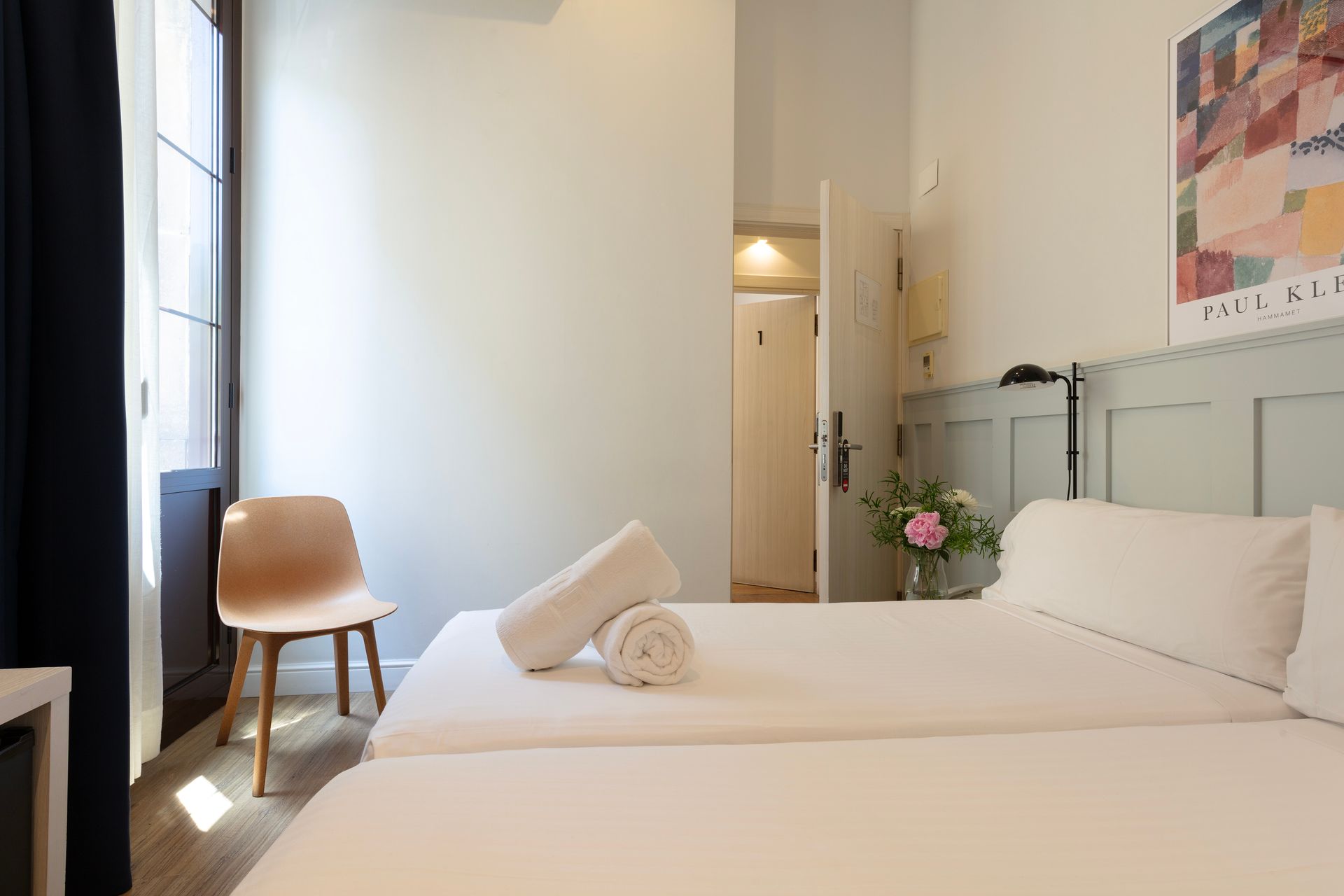Habitación luminosa con dos camas individuales, una silla de madera y un jarrón con flores junto a la ventana.
