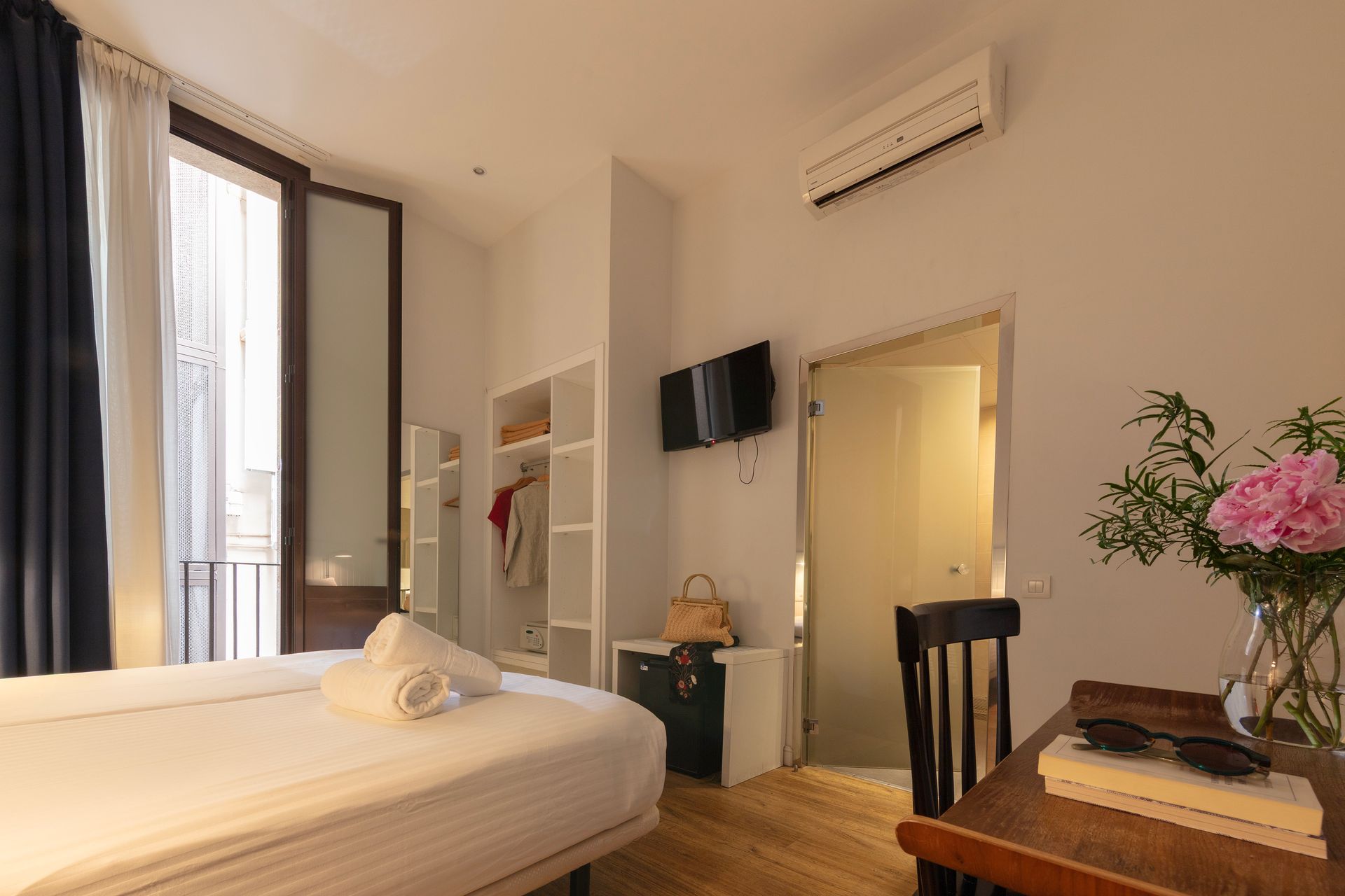 Habitación luminosa y moderna con cama, puerta de balcón, televisor de pared, aire acondicionado y una pequeña mesa.