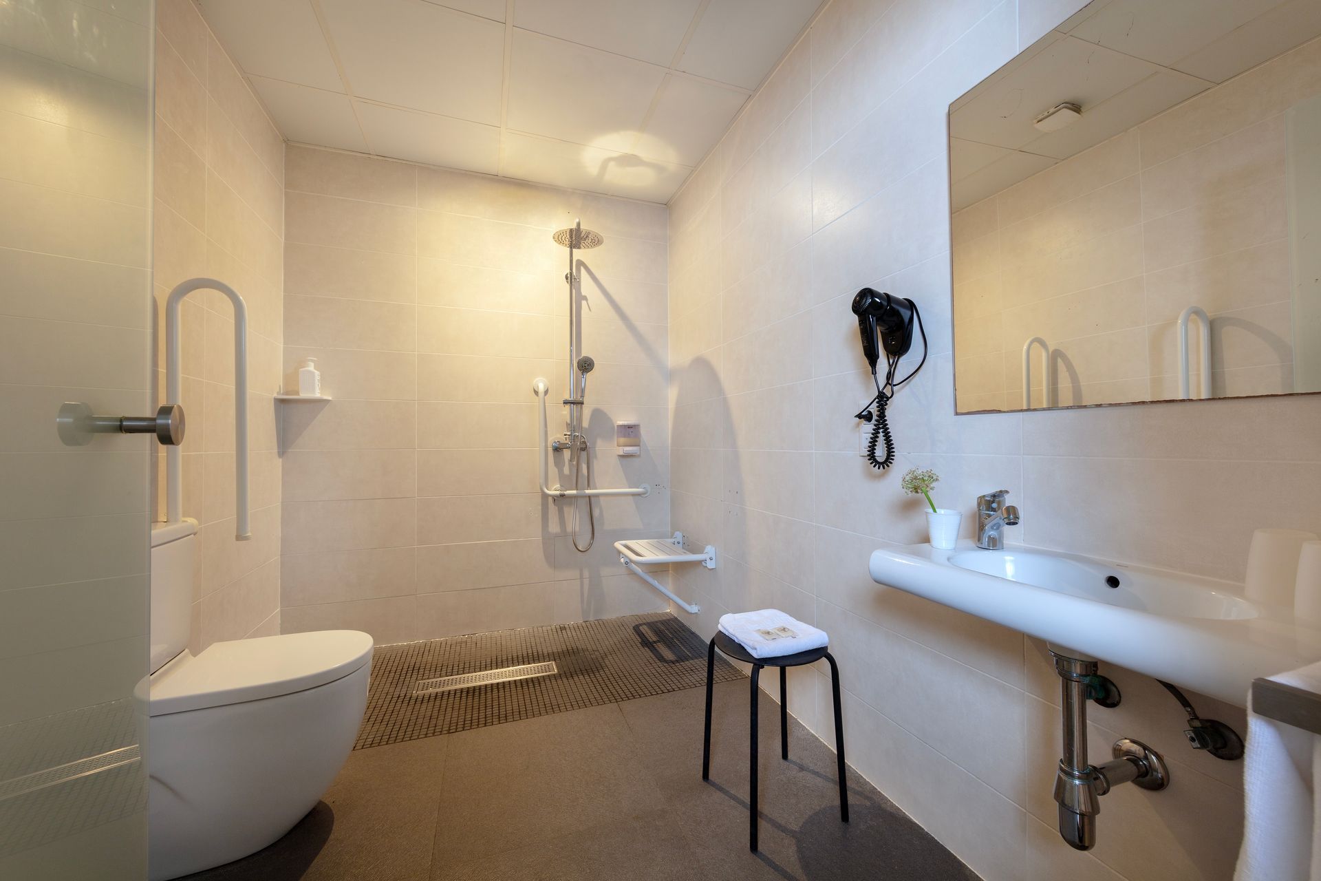 Baño accesible con inodoro, ducha, lavabo, barras de apoyo y taburete en una habitación blanca y luminosa.