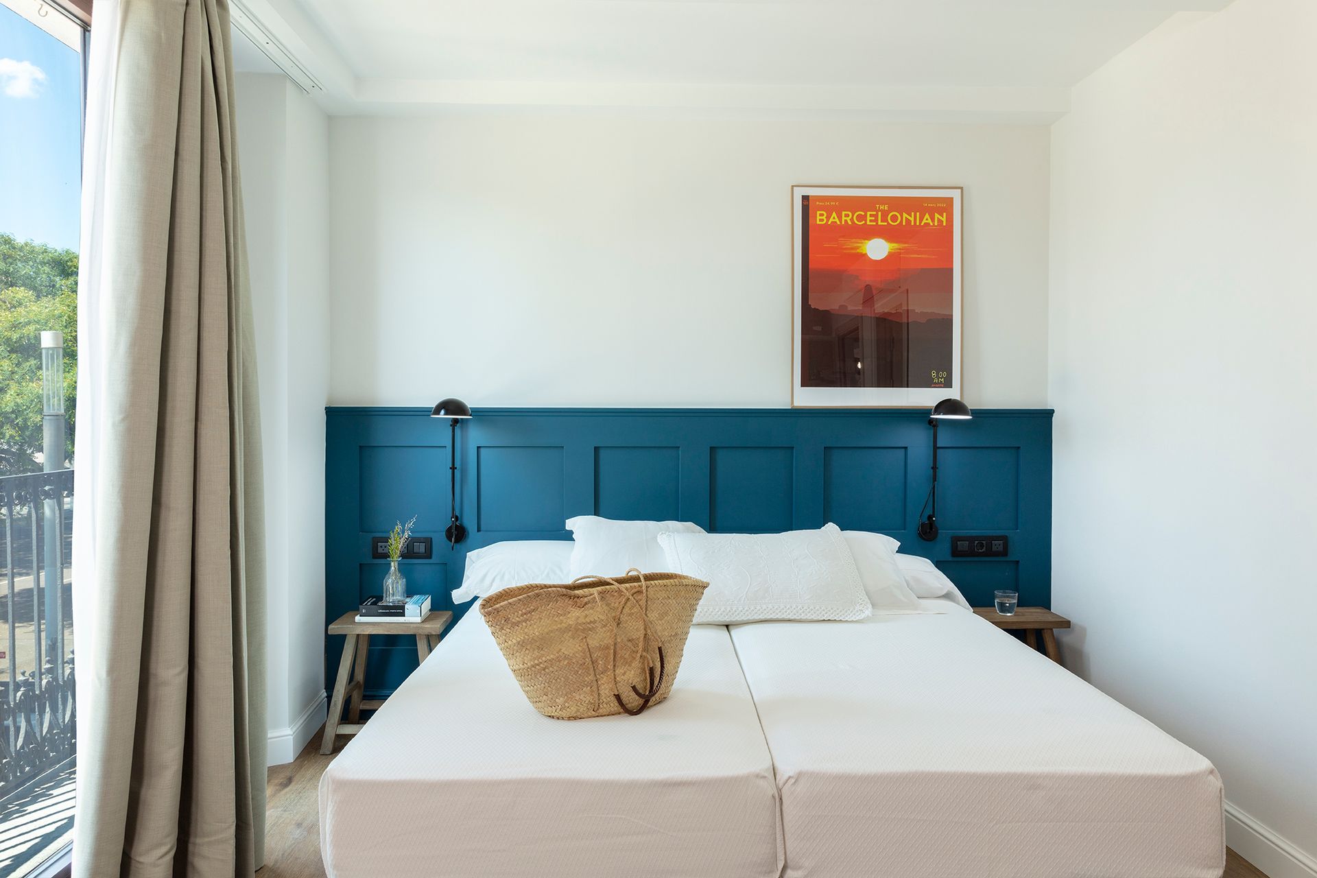Habitación luminosa con cabecero azul, ropa de cama blanca y una lámina enmarcada con una puesta de sol junto a la ventana.