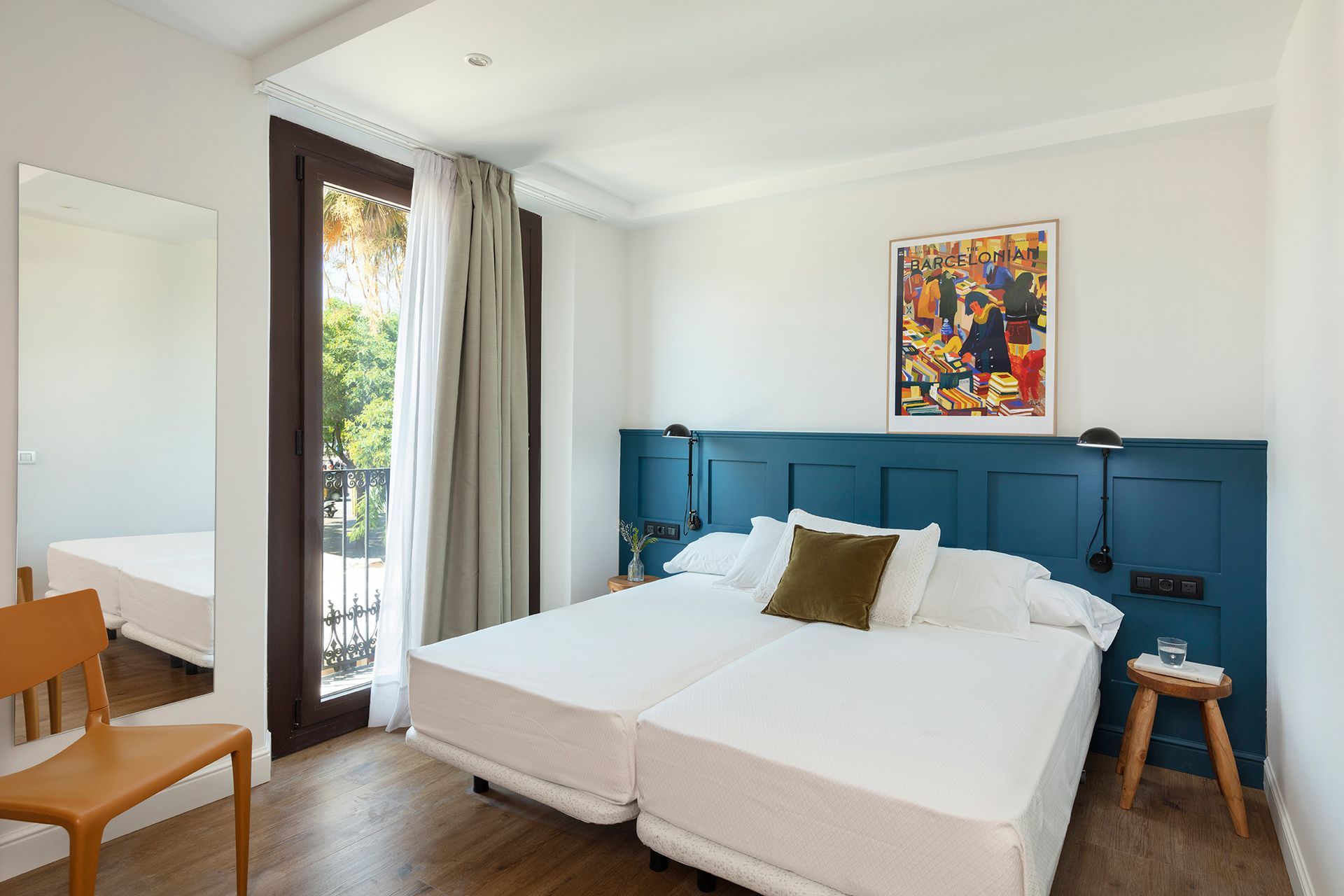 Habitación luminosa con cabecero azul, ropa de cama blanca, obras de arte y puerta de balcón abierta.
