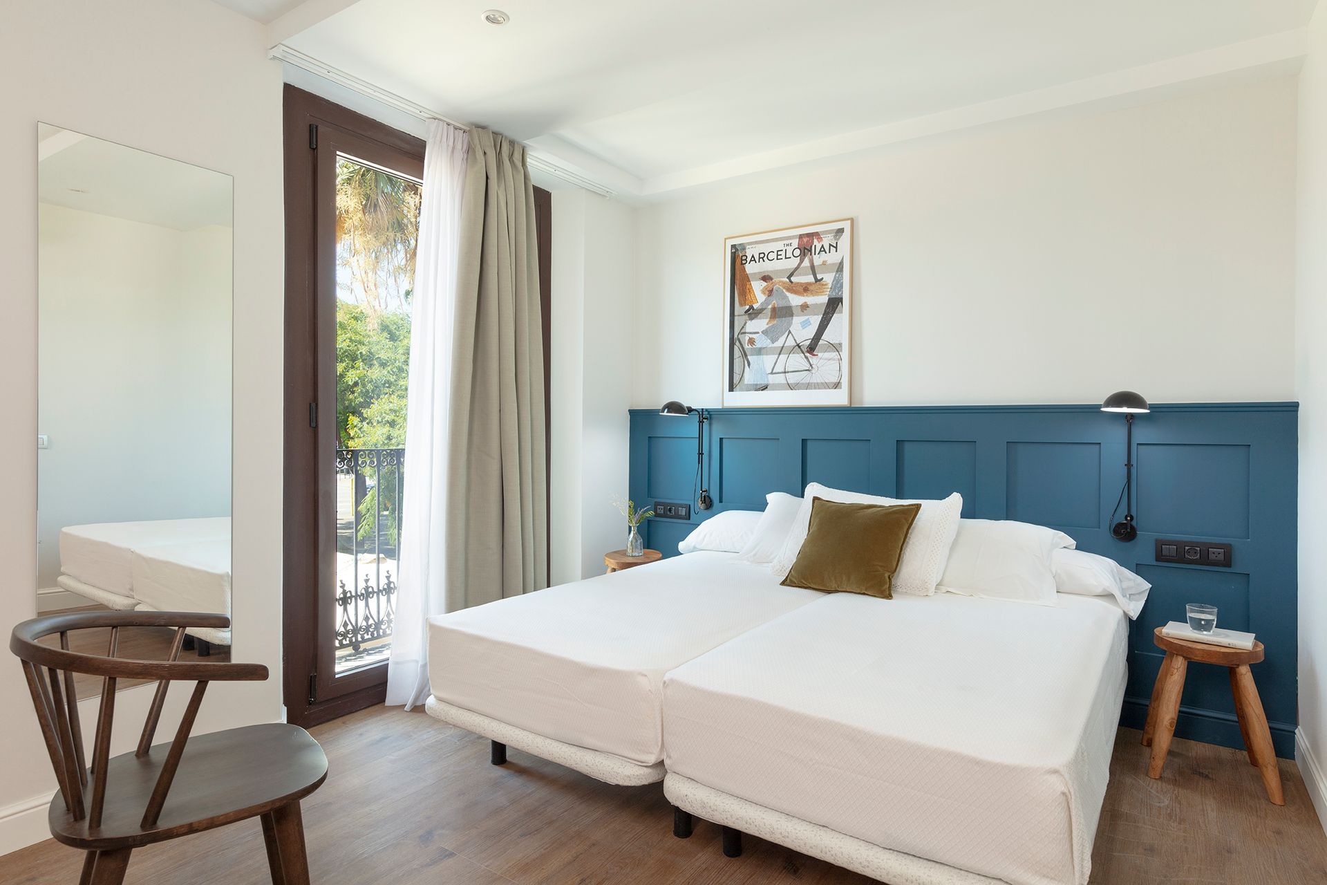 Habitación luminosa con cabecero azul, cama blanca, puerta que da al balcón y un pequeño taburete junto a la ventana.