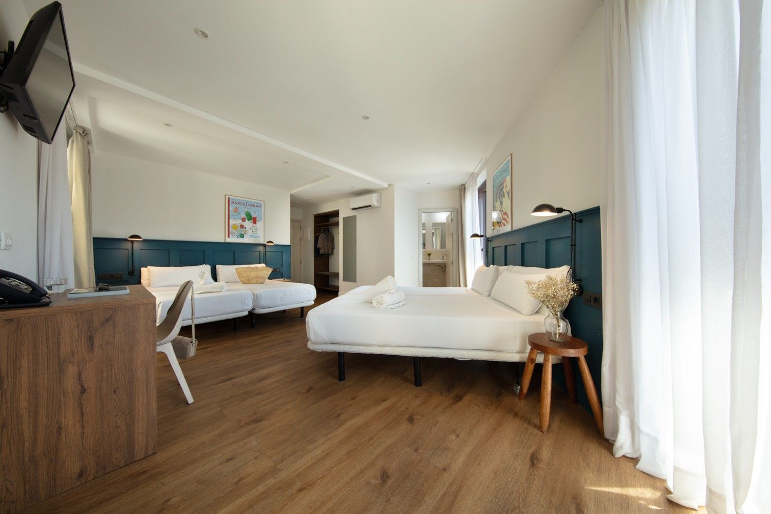 Una luminosa habitación de hotel con suelo de madera, dos camas con sábanas blancas, cabeceros azul oscuro, un  escritorio.