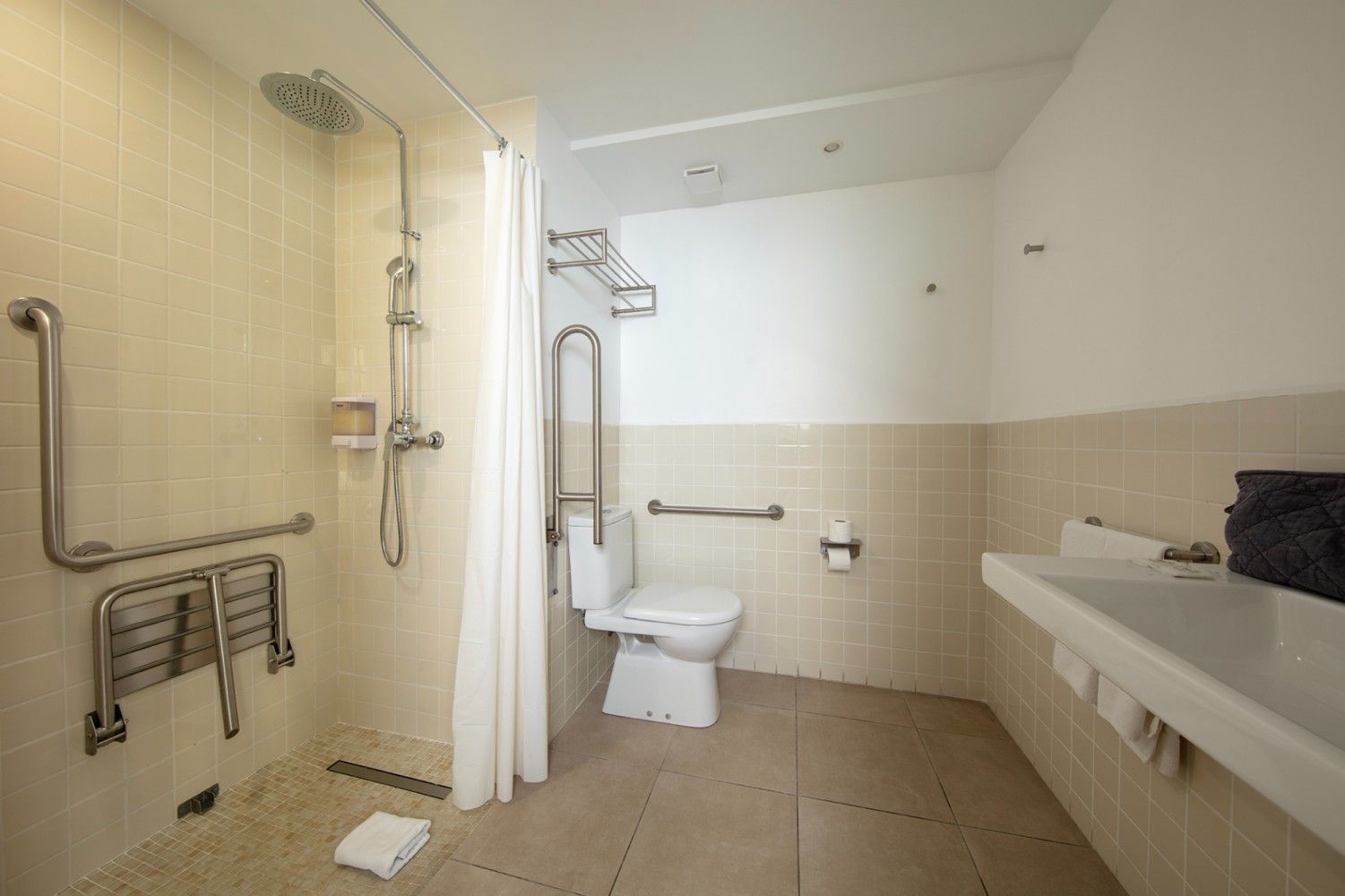 Baño accesible con ducha, inodoro, barras de apoyo y lavabo en un interior de azulejos beige.