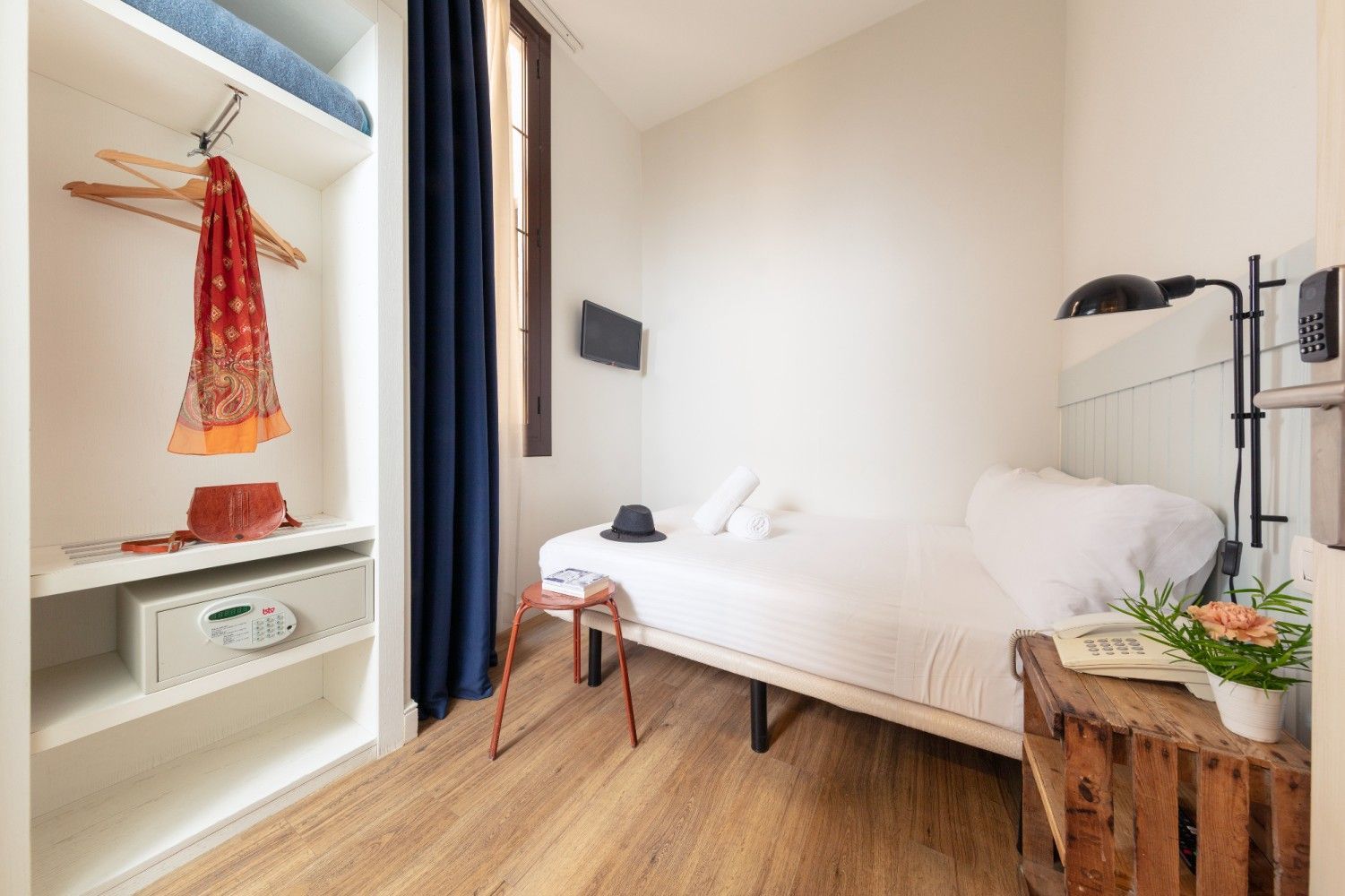 Una pequeña habitación de hotel con una cama individual, una mesita de noche de madera, una lámpara de pared.