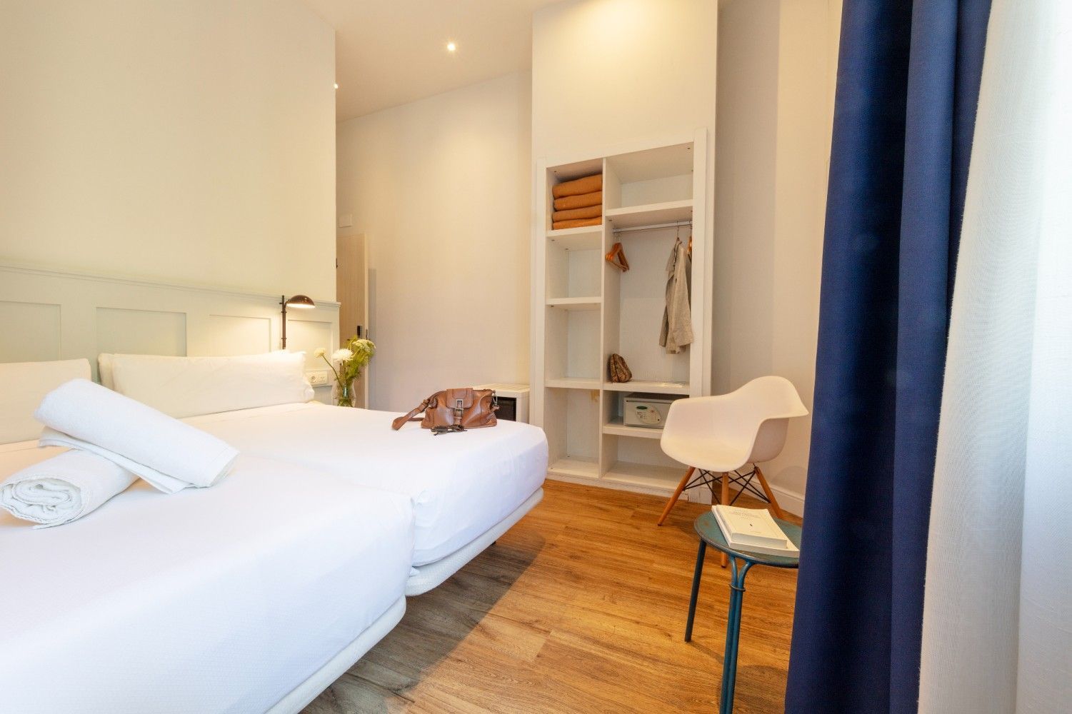 Dormitorio minimalista con cama blanca, suelo de madera, armario abierto y cortinas azules.