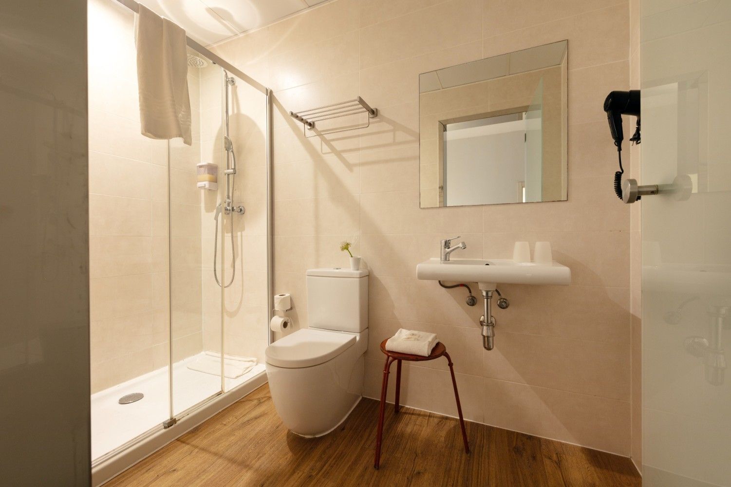 Un baño moderno con ducha con mampara de cristal, inodoro blanco, lavabo de pared, espejo y taburete sobre suelo de madera.