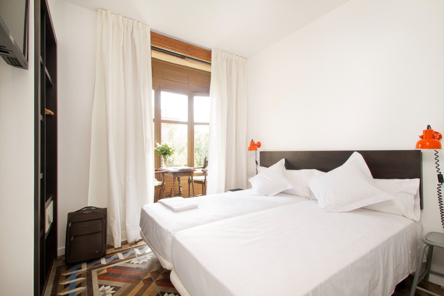 Habitación luminosa con sábanas blancas, ventana alta, cortinas transparentes y alfombra estampada.
