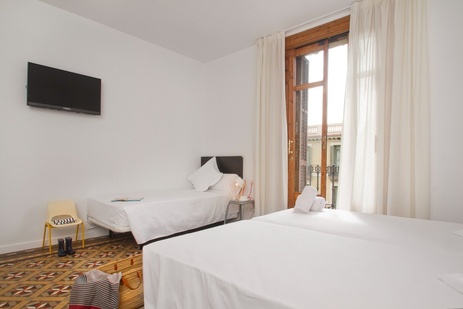 Habitación de hotel luminosa con dos camas individuales, televisor de pared, alfombra estampada y ventana alta con cortinas.