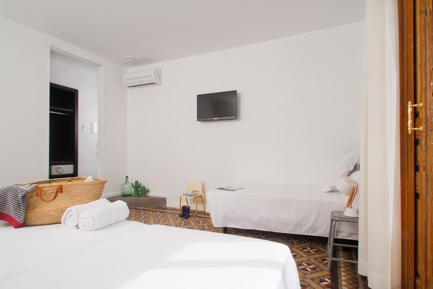 Habitación de hotel luminosa con dos camas, televisor montado en la pared y silla de mimbre junto a la ventana.