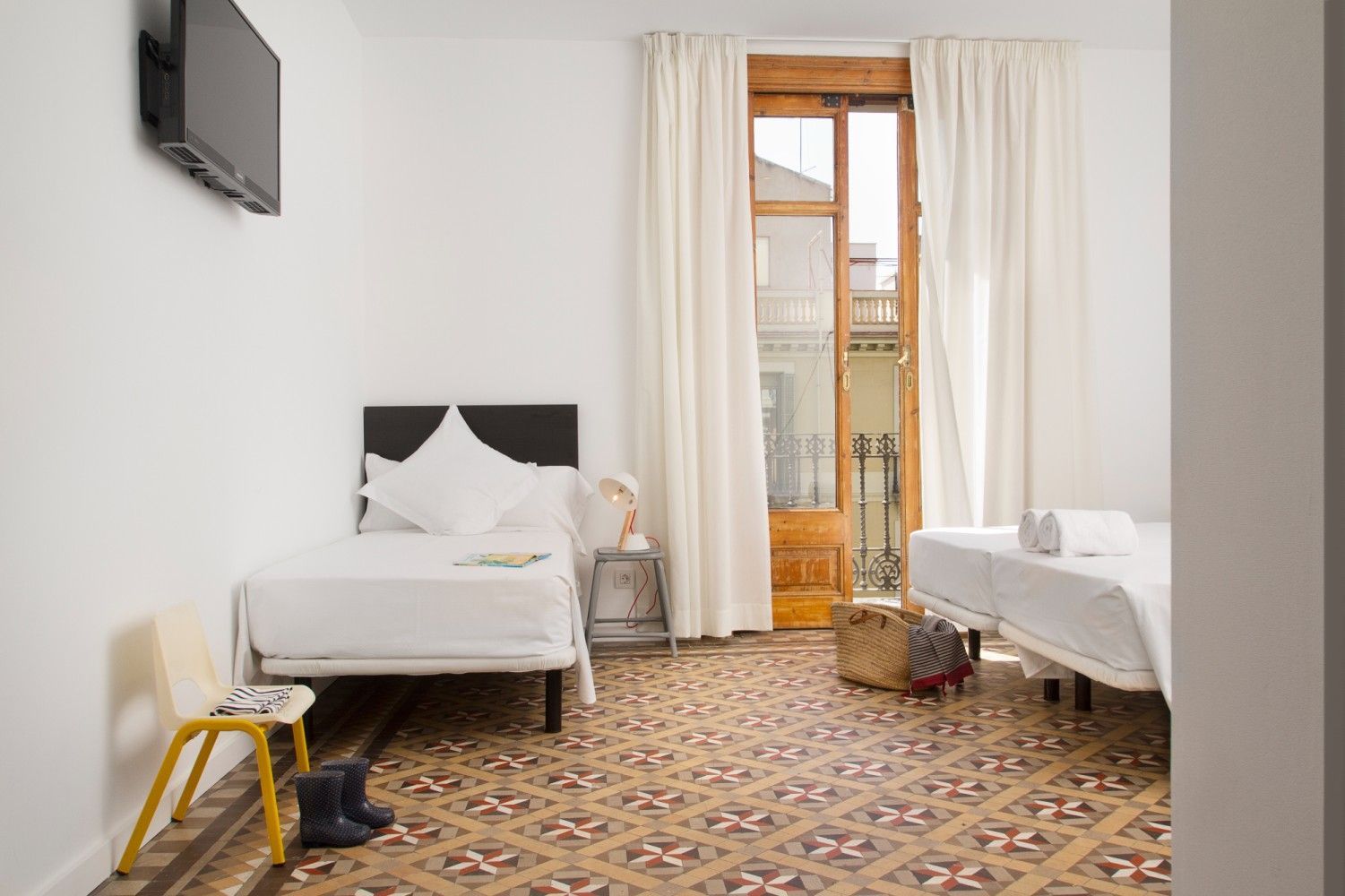 Una pequeña habitación con dos camas individuales, alfombra estampada y puerta de balcón con cortinas blancas.