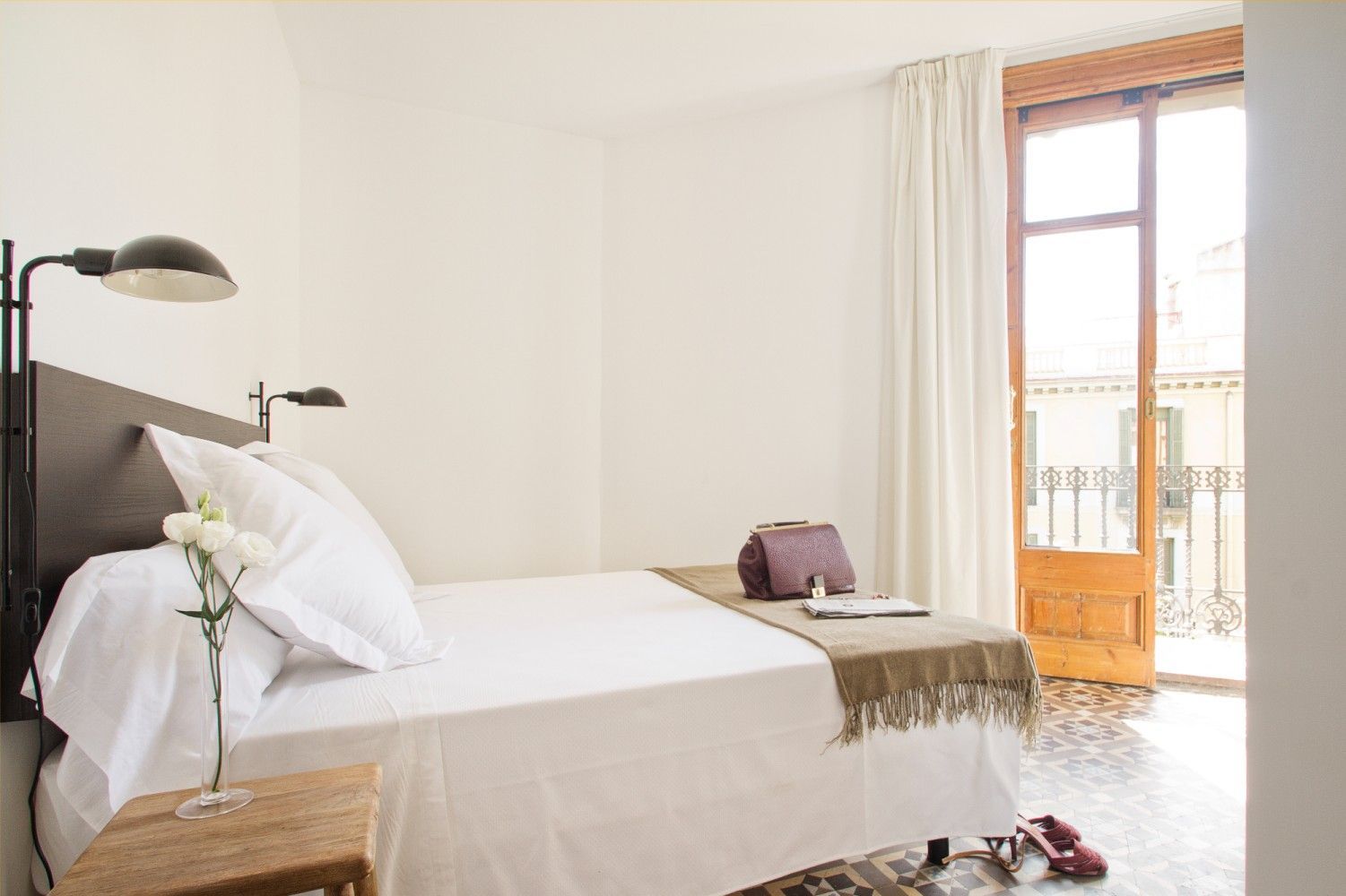 Habitación luminosa con una cama bien hecha, lámpara de noche y puerta de balcón abierta con cortinas.