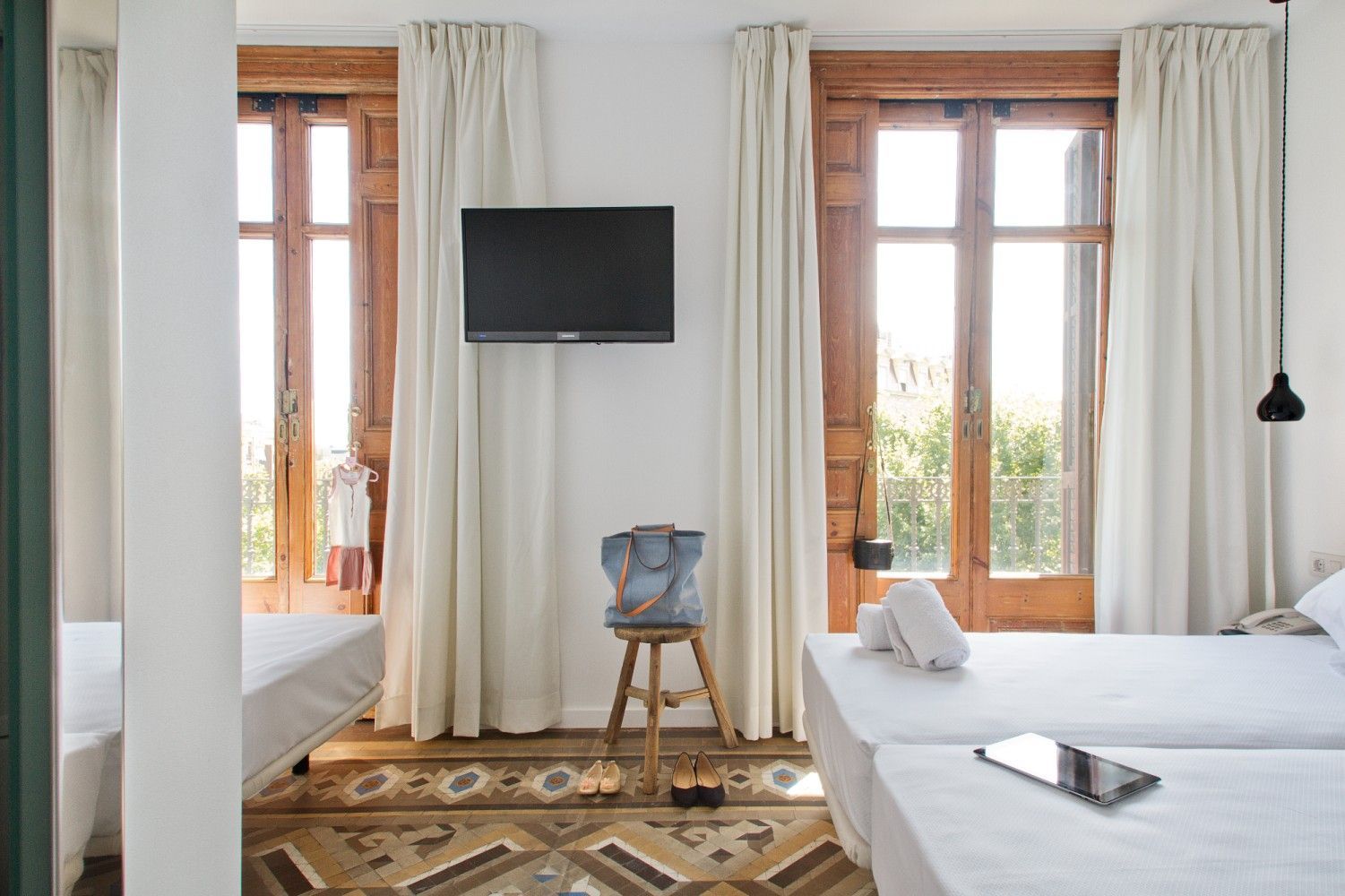 Habitación luminosa con dos camas individuales, televisor montado en la pared y grandes ventanas con cortinas blancas.