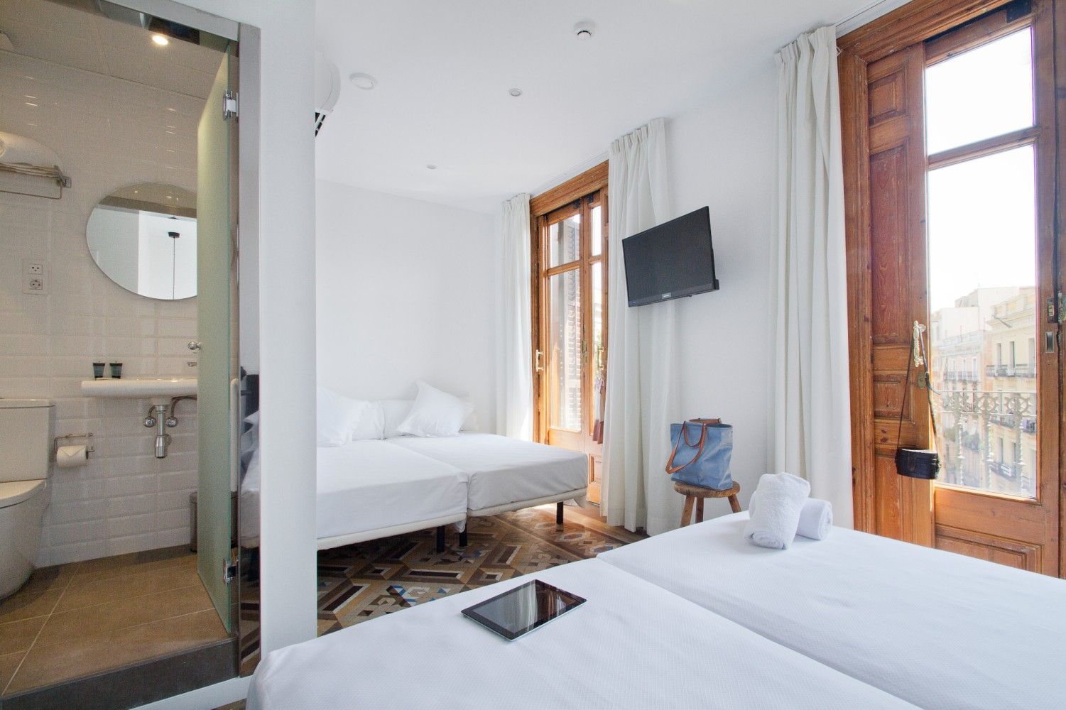 Habitación de hotel luminosa con dos camas, televisor de pared, baño y grandes puertas de madera con vistas a la ciudad.