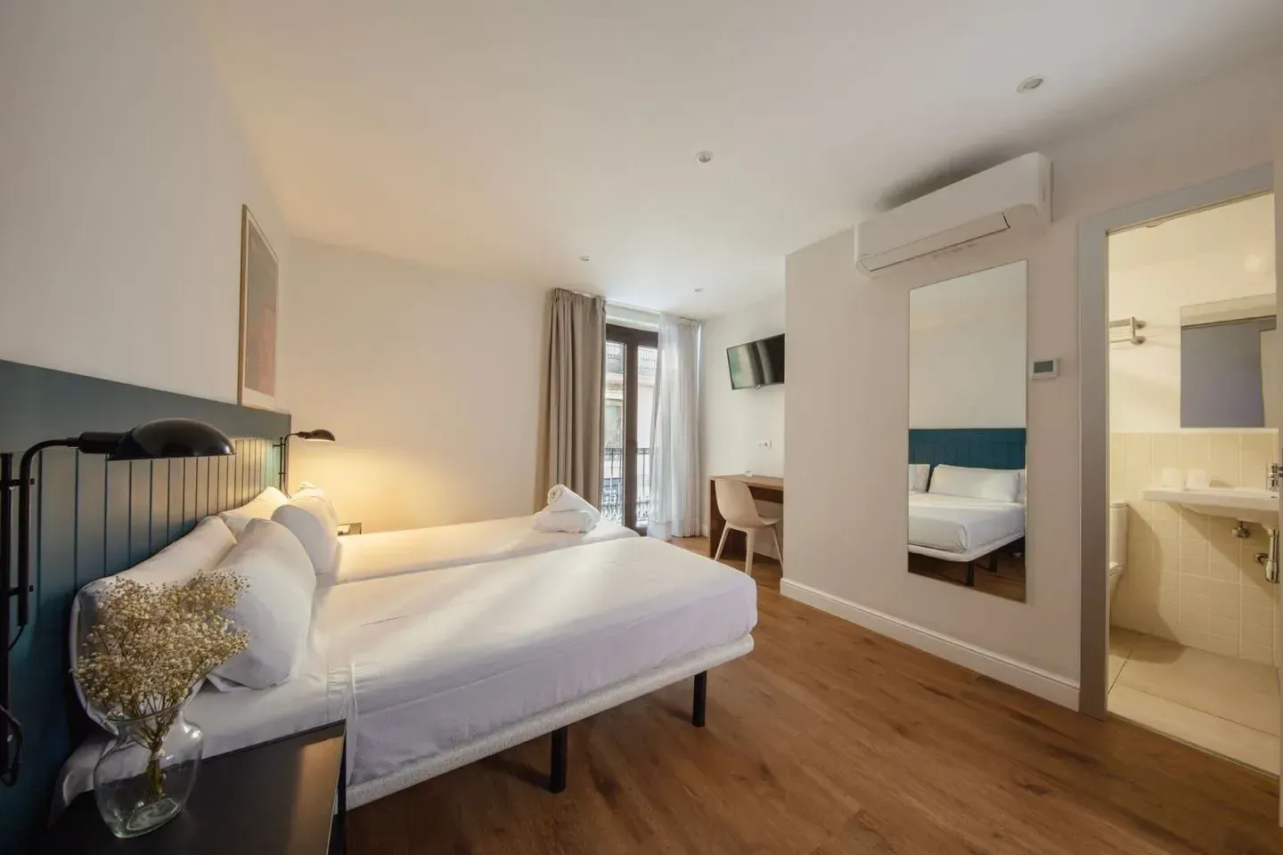 Una habitación de hotel moderna y bien iluminada con dos camas blancas, pisos de madera, un espejo y un baño contiguo.