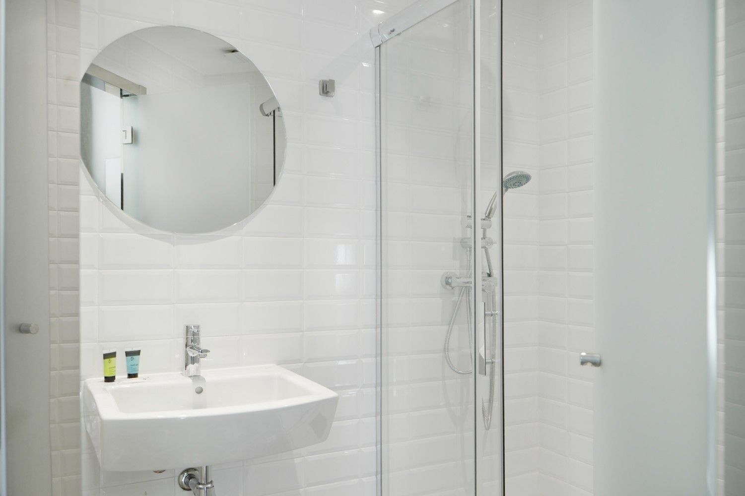 Baño moderno de color blanco brillante con lavabo de pared, espejo redondo y cabina de ducha de cristal.