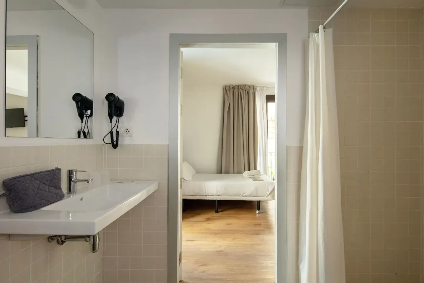 Un baño moderno con lavabo y espejo blancos, que se abre a un dormitorio luminoso con cama y cortinas de colores claros.